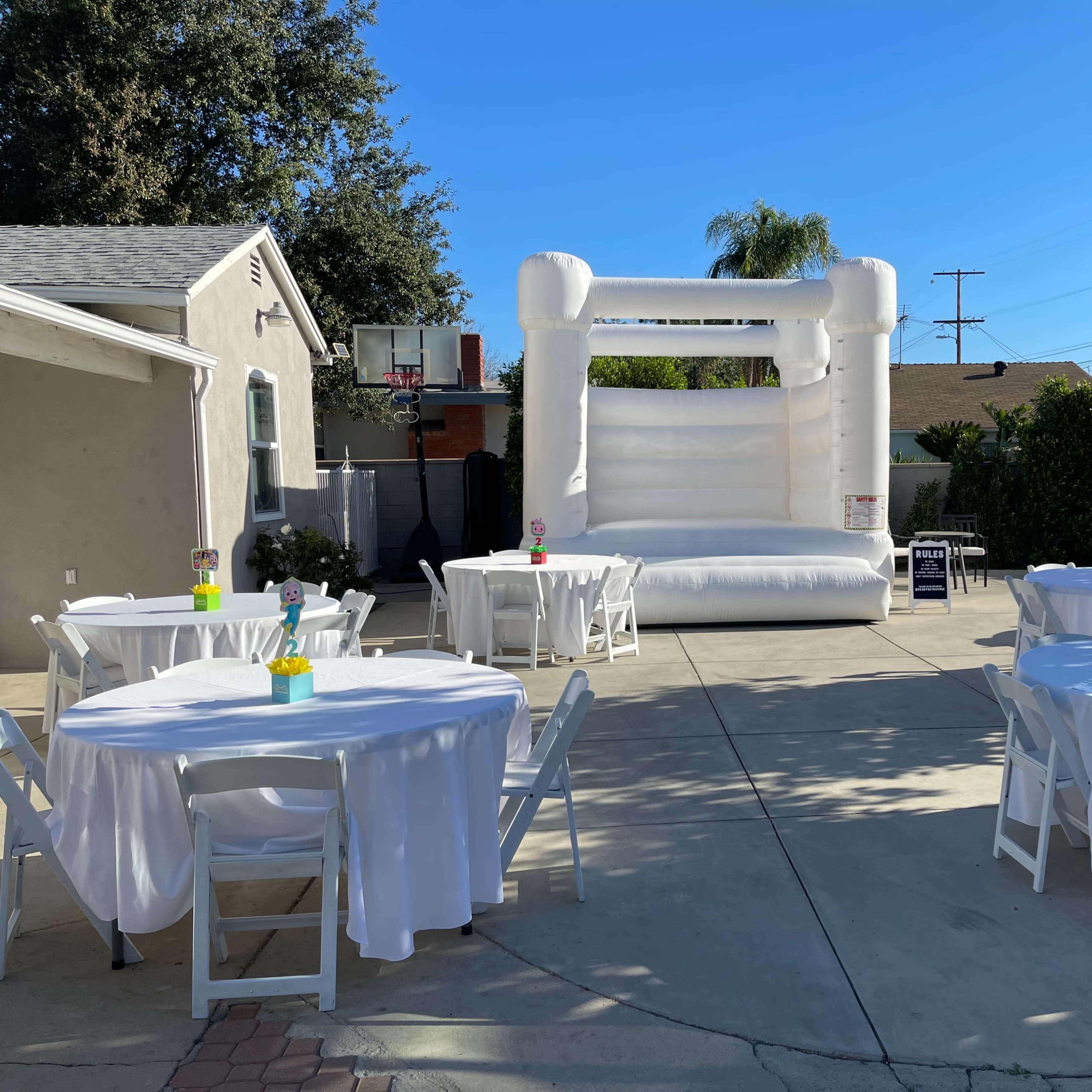 A backyard setting features a white bouncy castle and several round tables covered with white tablecloths.
