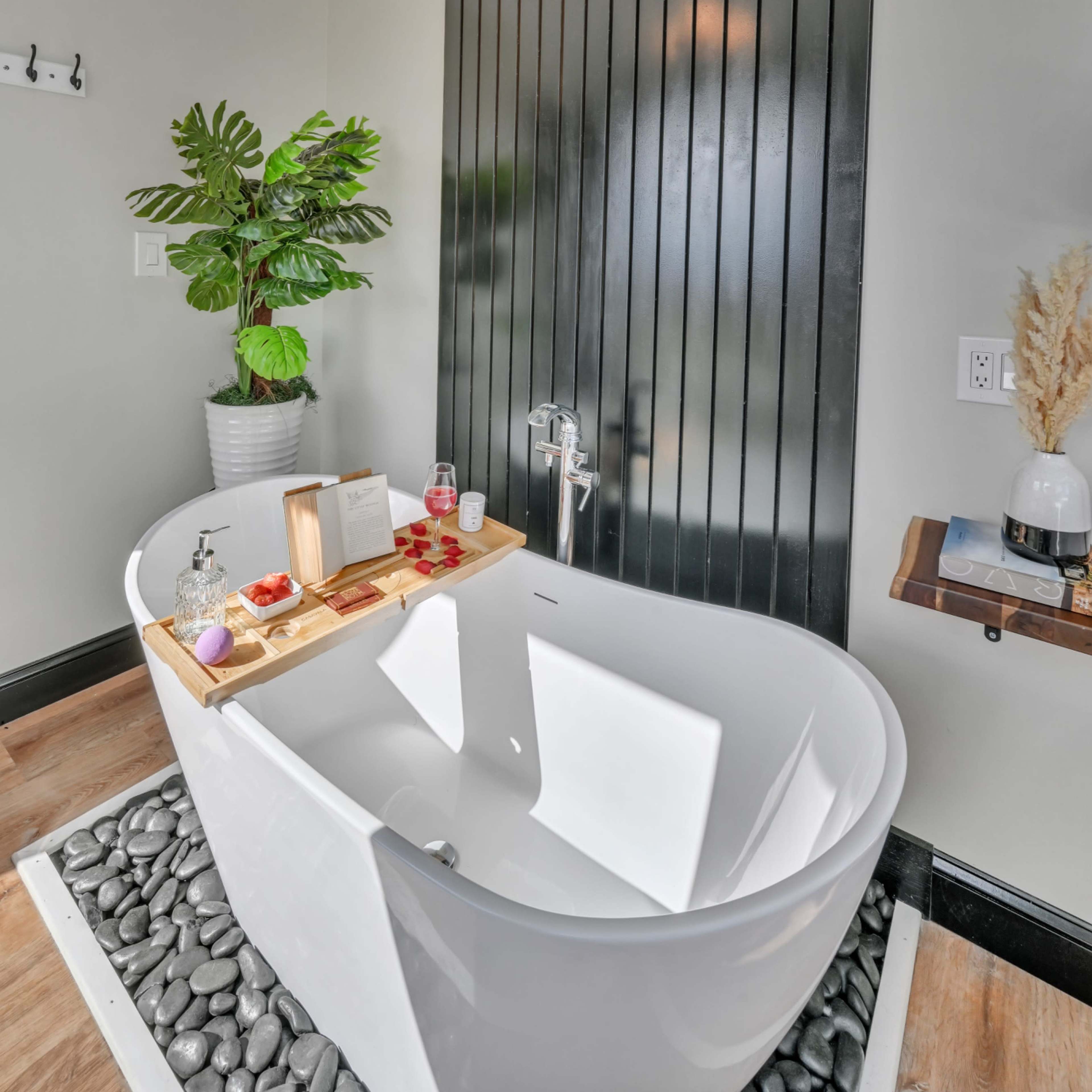 A modern bathroom features a freestanding white bathtub, a potted plant, and a decorative wooden tray on the tub's edge.