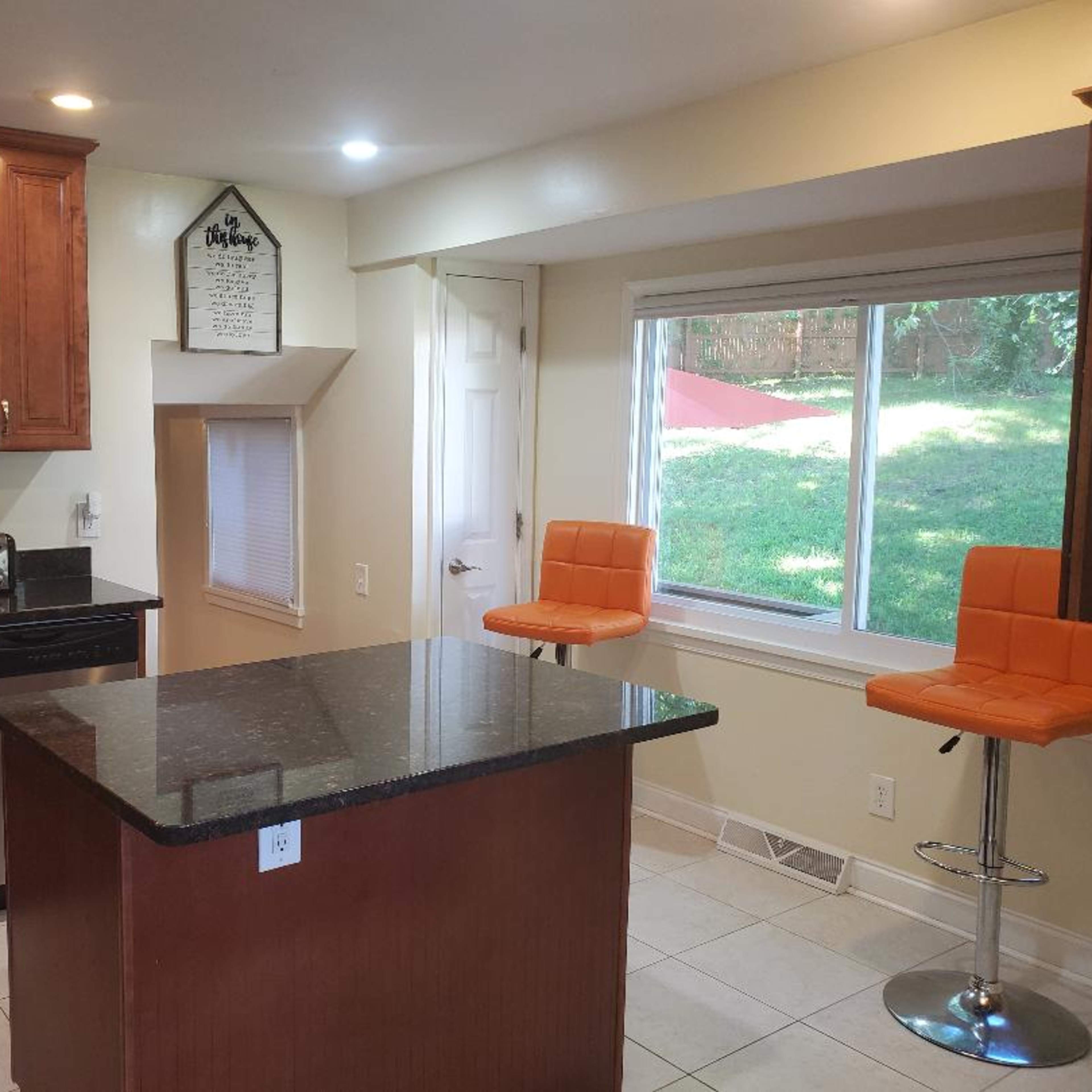 A kitchen with wooden cabinets, a black countertop island, two orange bar stools, and a window overlooking a grassy area.