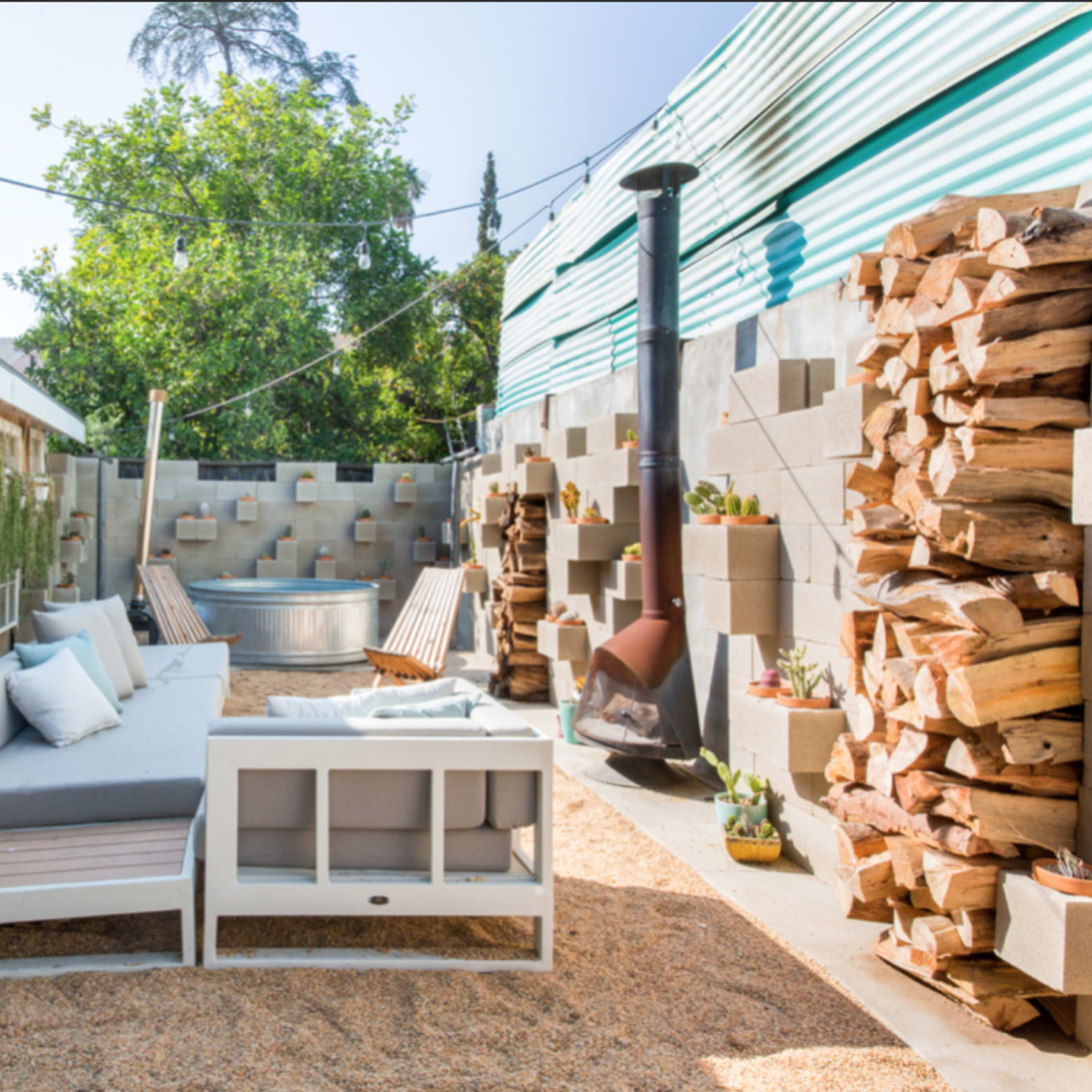 The scene features a garden area with a wooden furniture set, a metal tub, stacked firewood, and plants, surrounded by a wall and a building.