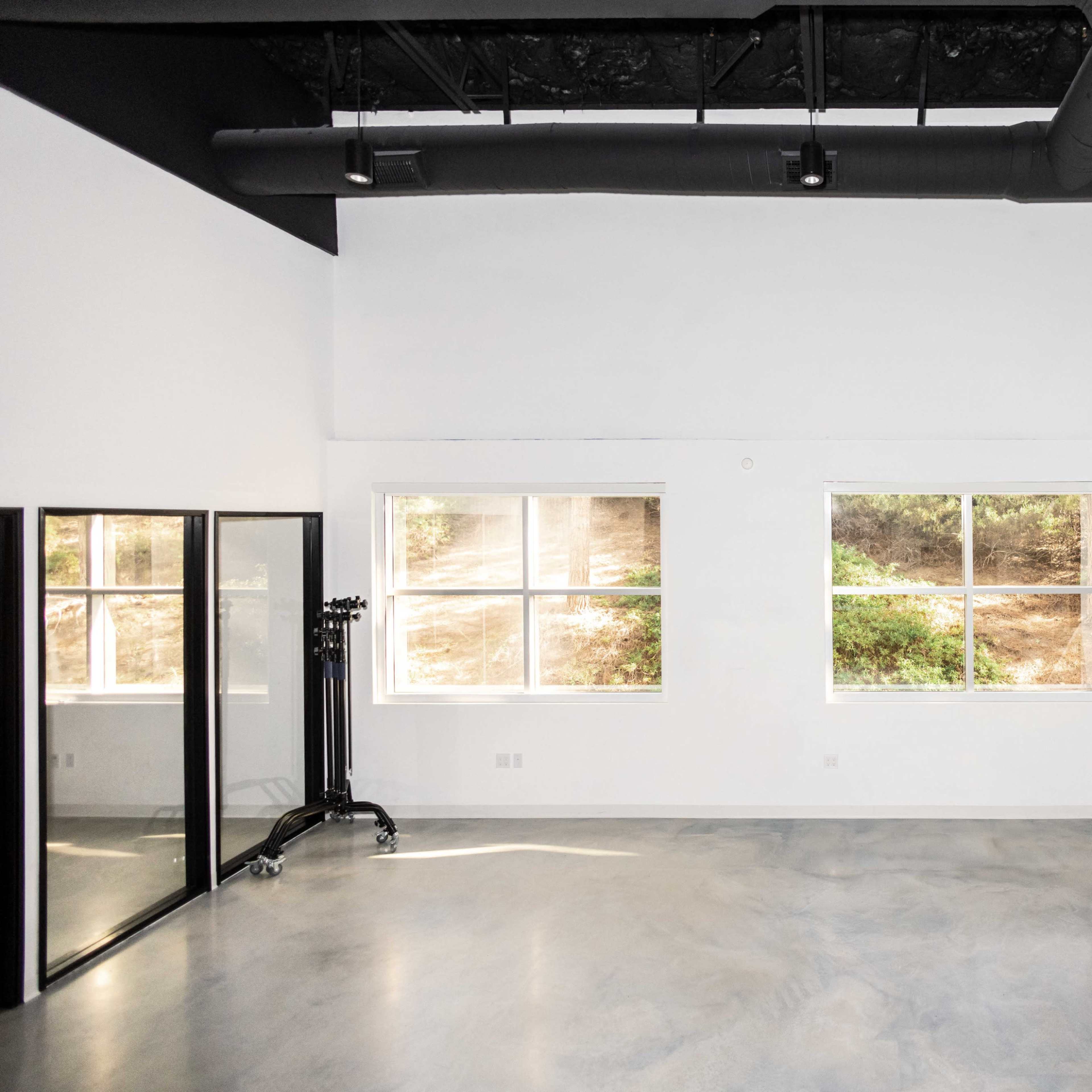 An empty room with large windows, a concrete floor, and minimal furnishings, featuring a mirror and a coat rack on one side.