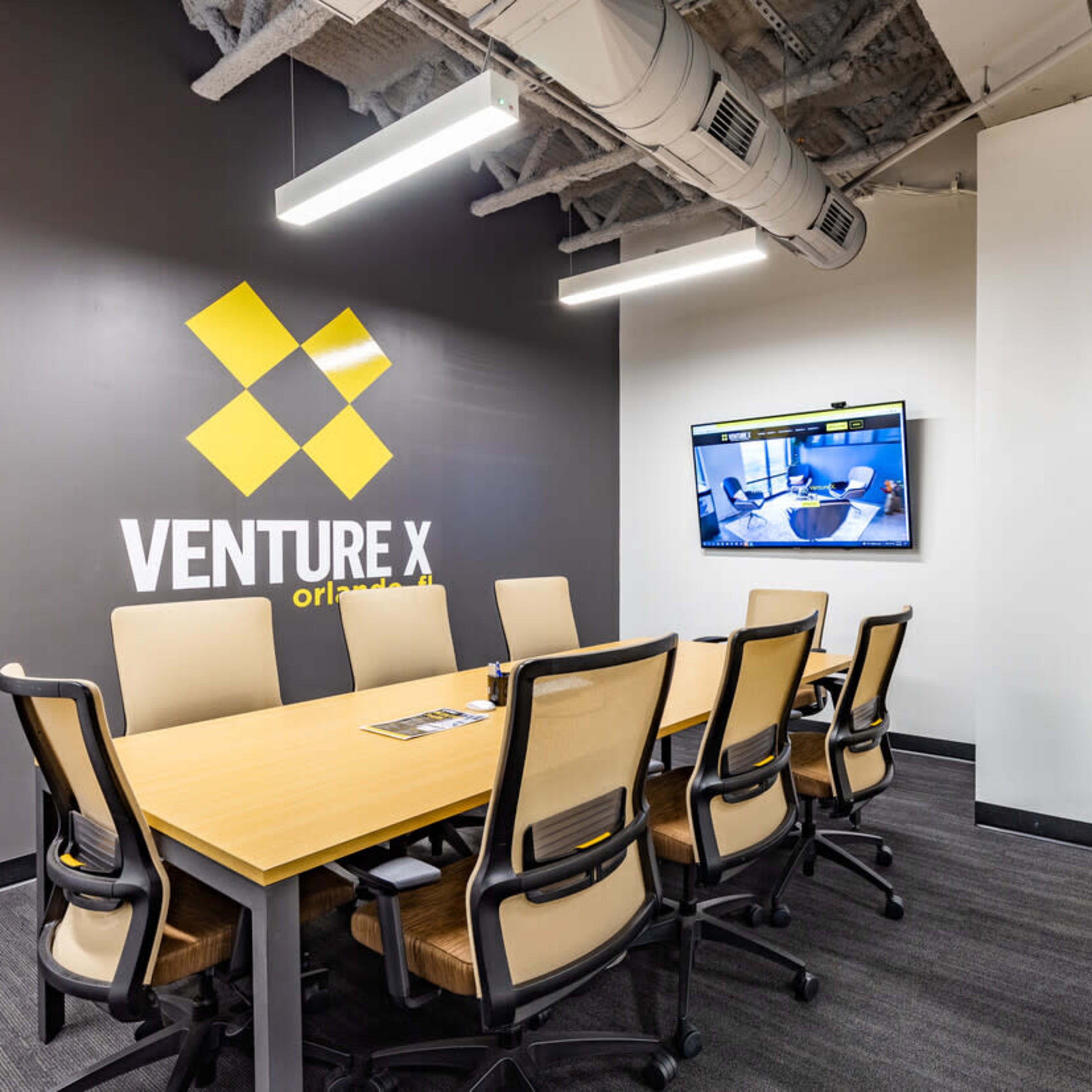 A modern conference room features a large table surrounded by eight chairs, along with a wall-mounted screen and a potted plant in the corner.
