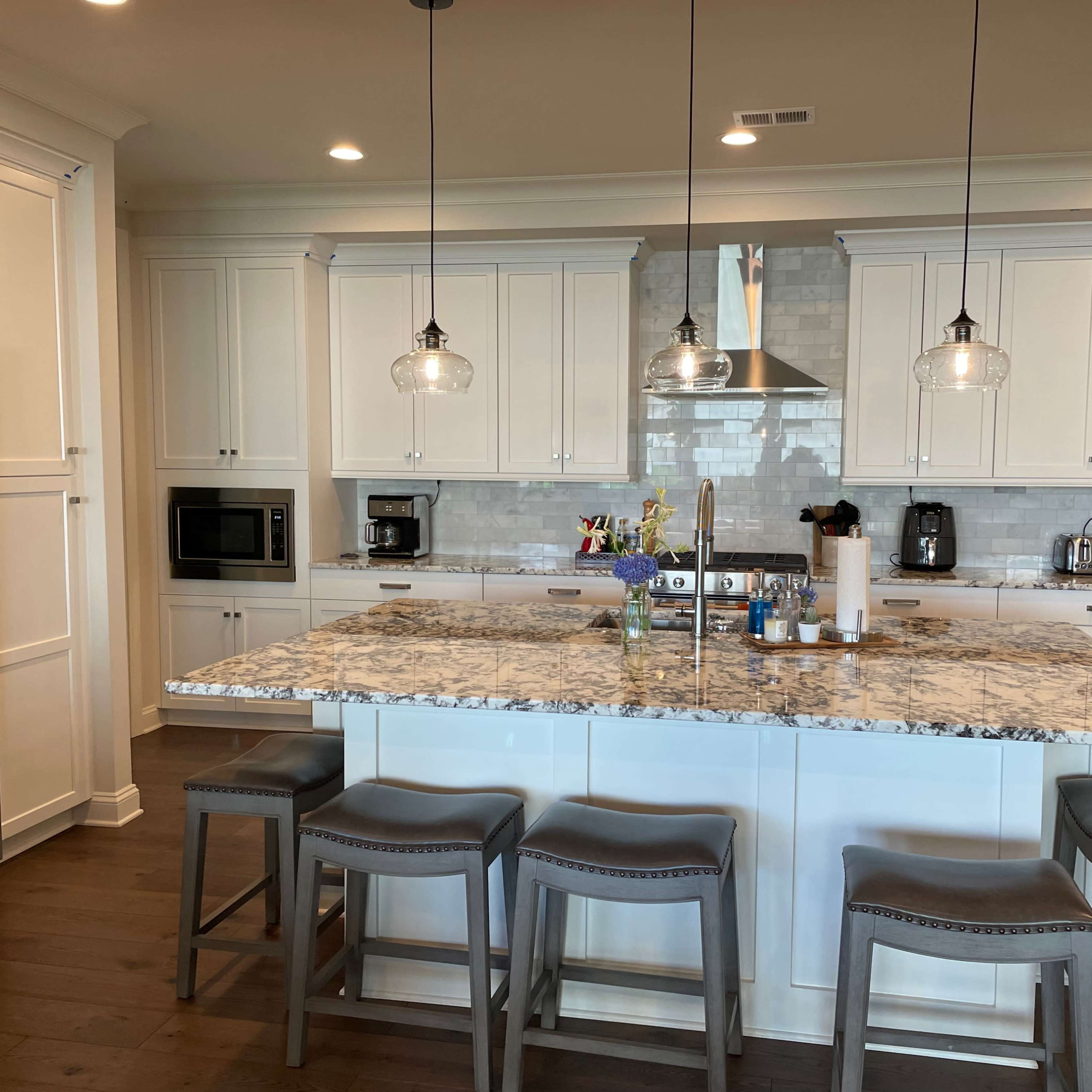 A modern kitchen with white cabinetry, a large granite island, and three grey bar stools arranged around it.