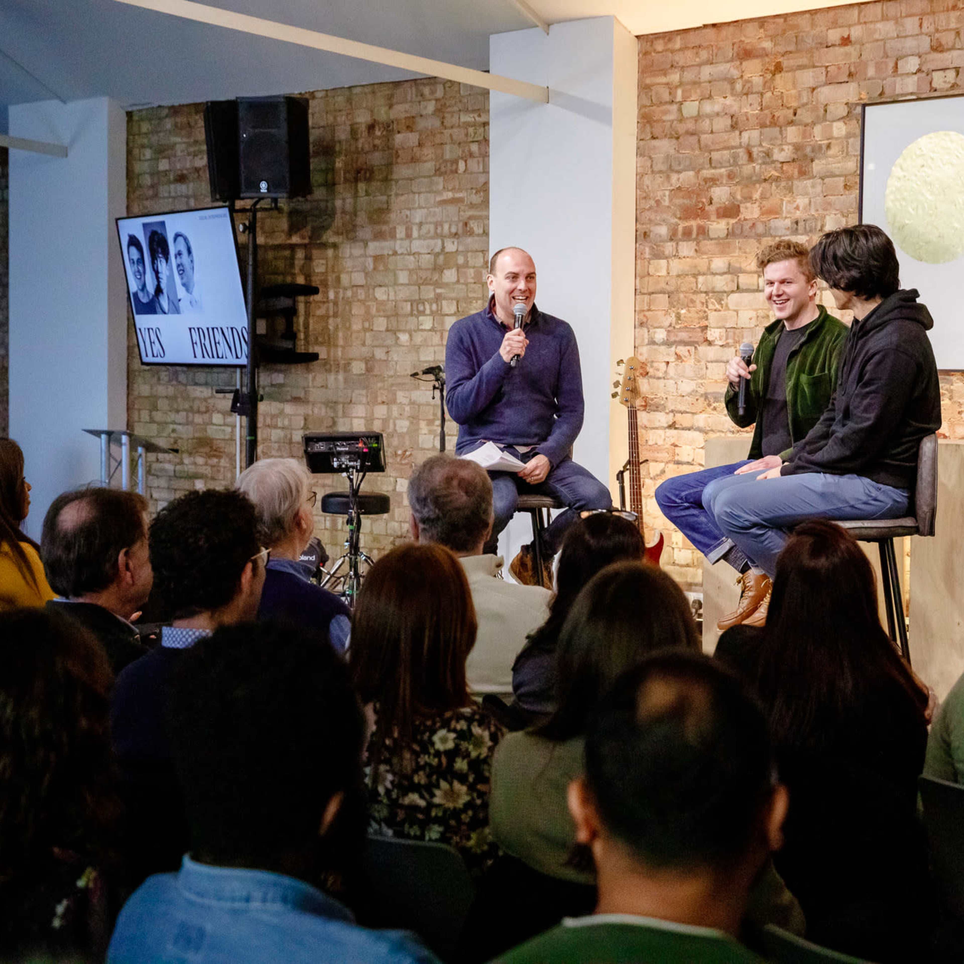 A panel discussion is taking place with three speakers seated on a stage in front of an audience in a brick-walled venue.