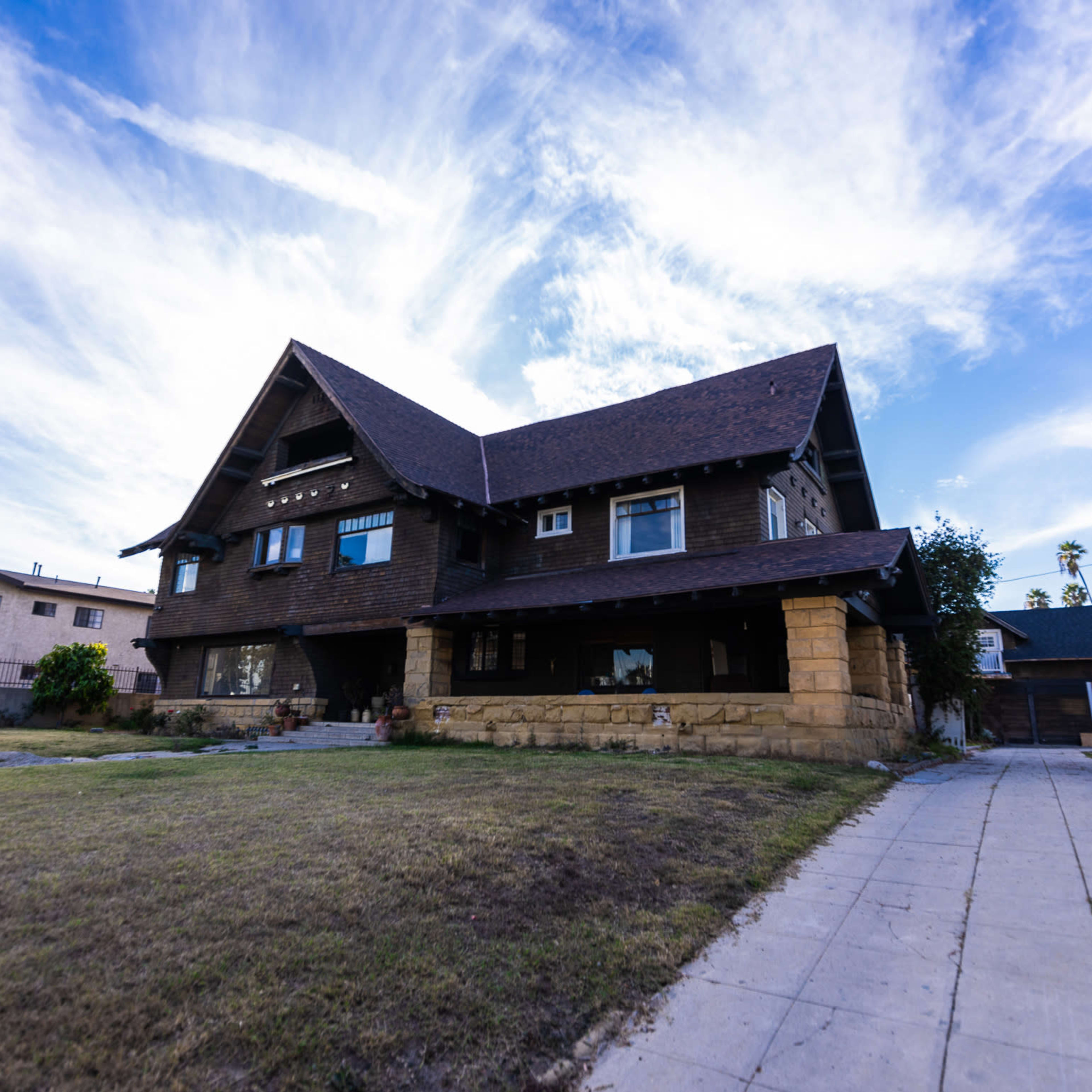 A large, two-story brown house with a steeply pitched roof and stone foundation is set against a blue sky with scattered clouds.