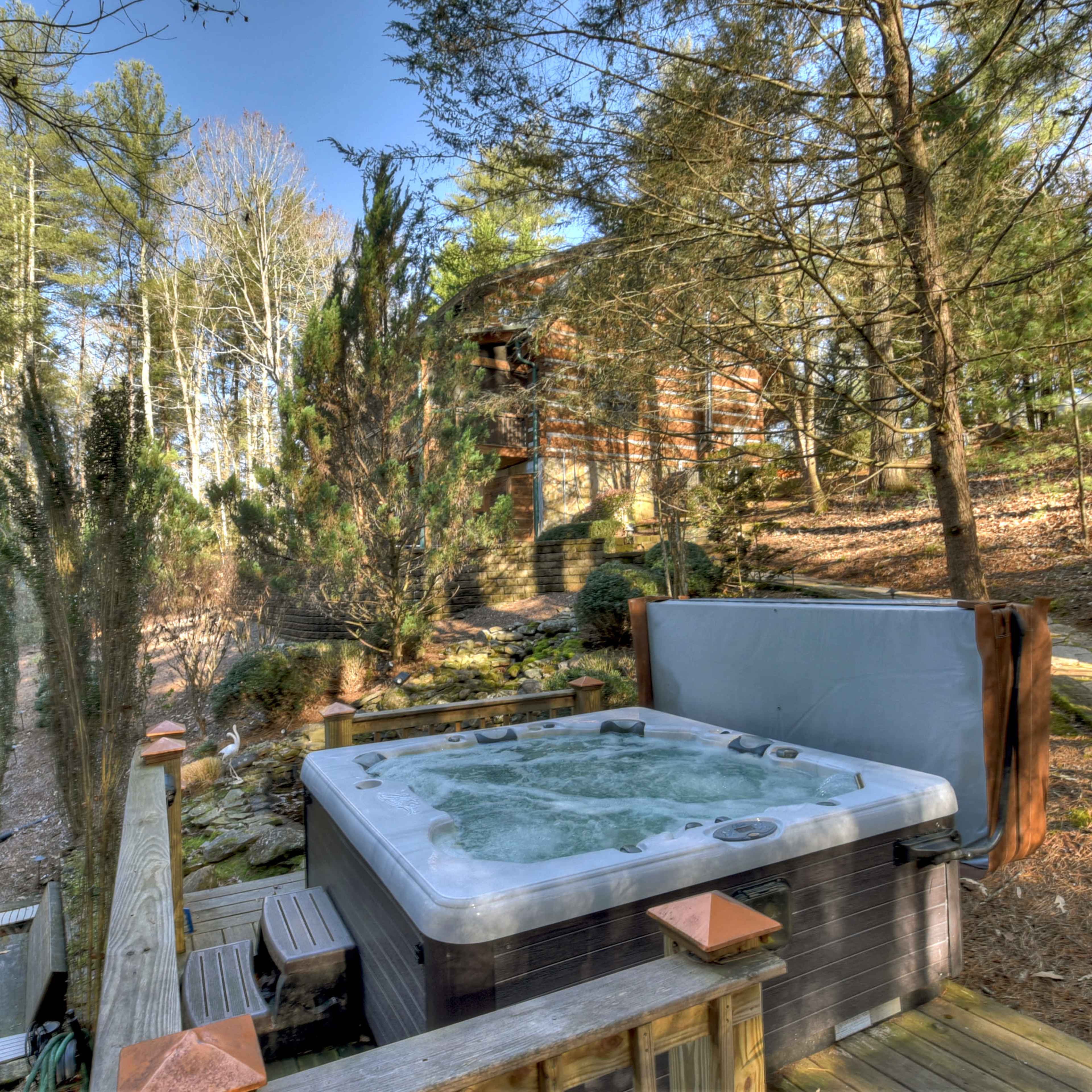 A hot tub sits on a wooden deck surrounded by trees and a pathway leading to a cabin.