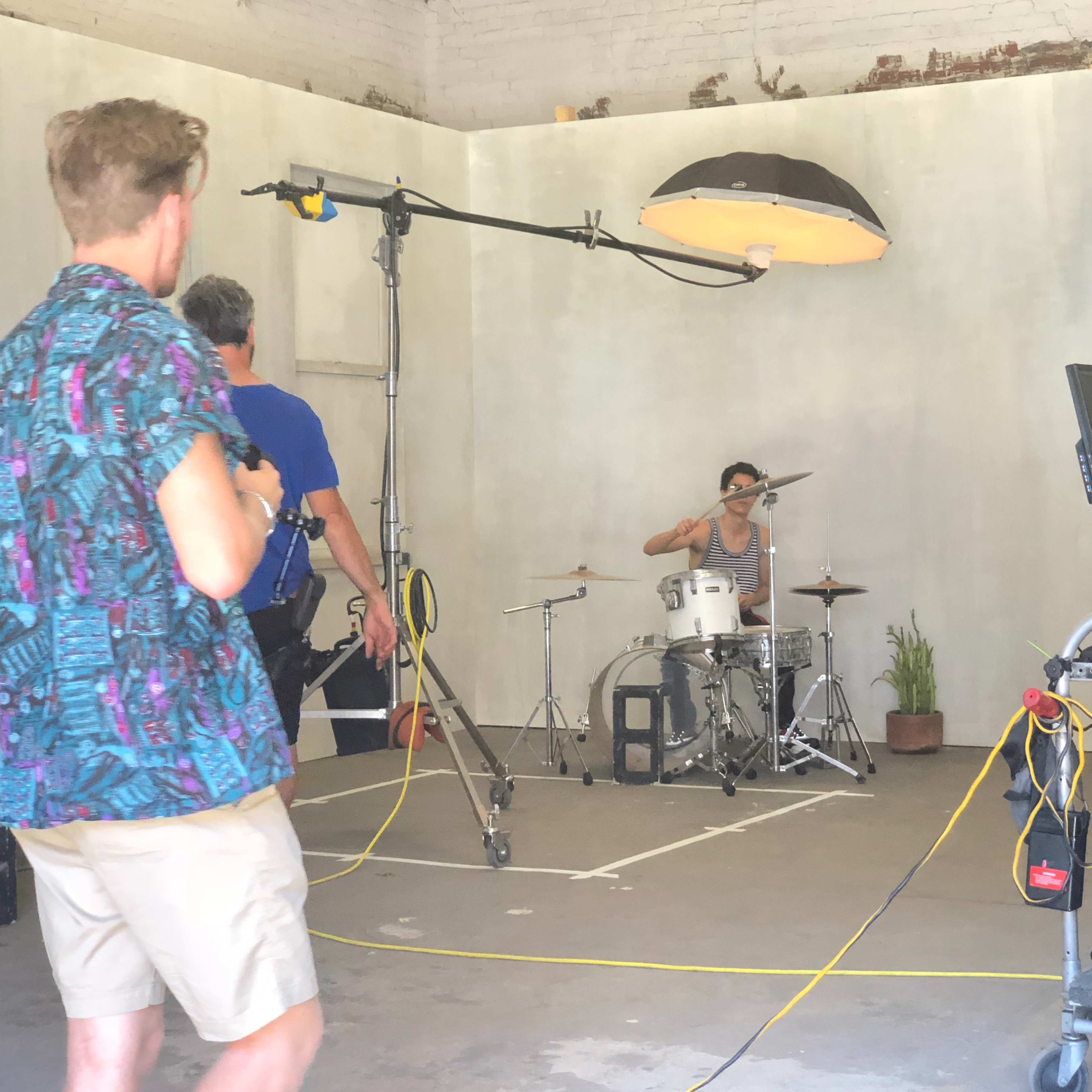 A drummer performs on a small set while a crew operates cameras and lighting equipment in an industrial space.