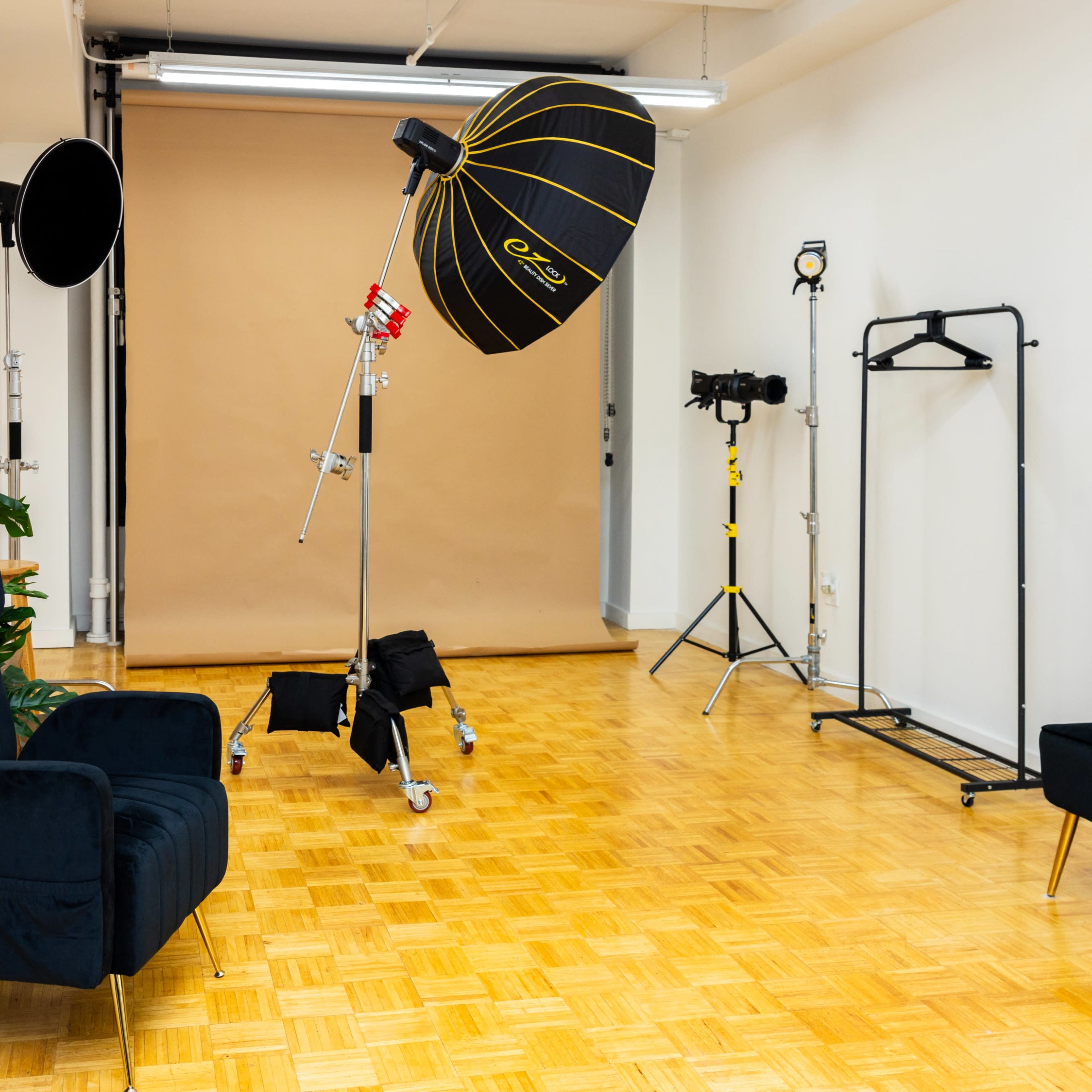 A photo studio with a light setup, two seating areas, a clothing rack, and a plain backdrop.