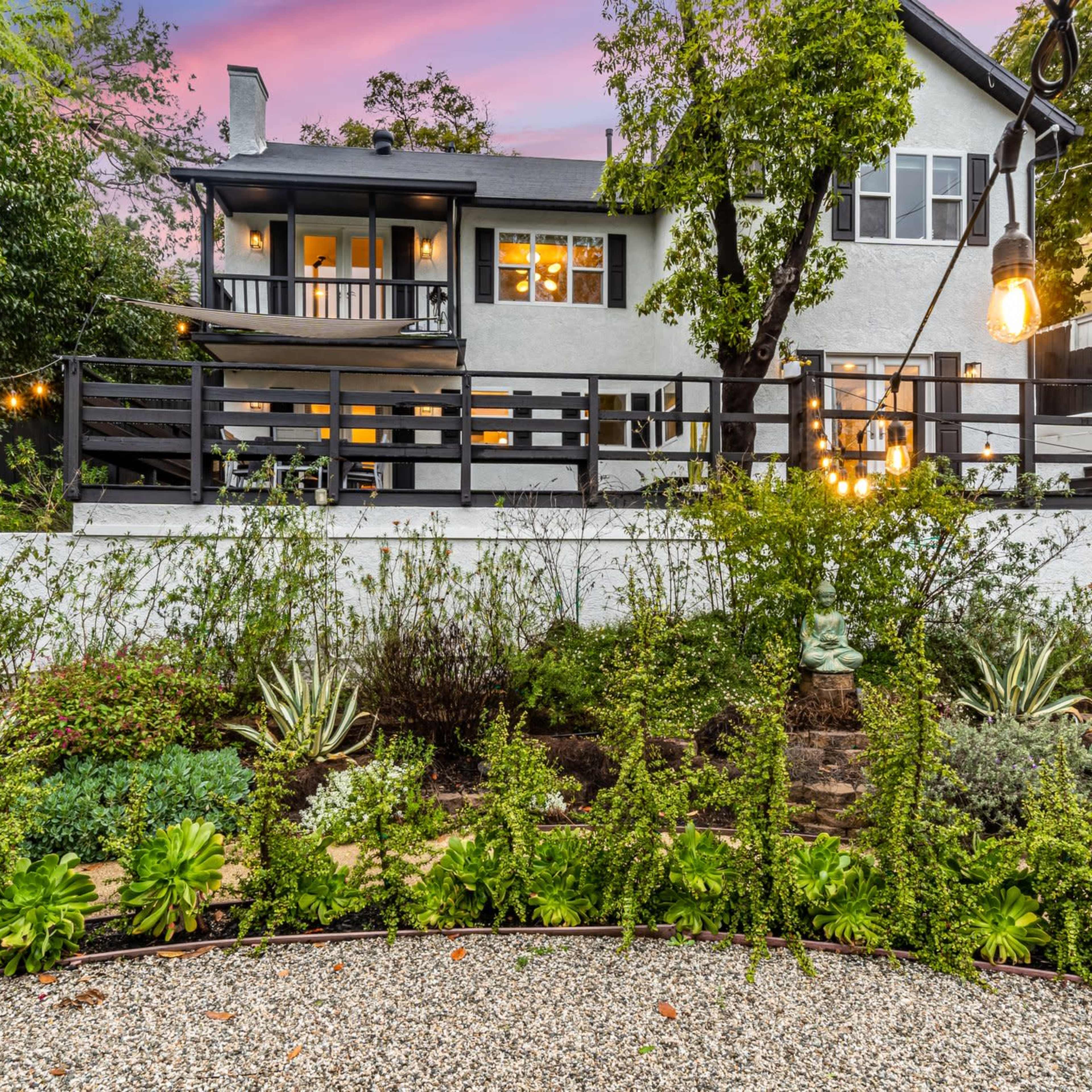 A well-kept garden with various plants and a circular stone pathway leads to a modern two-story house with a deck, surrounded by trees and illuminated by string lights.