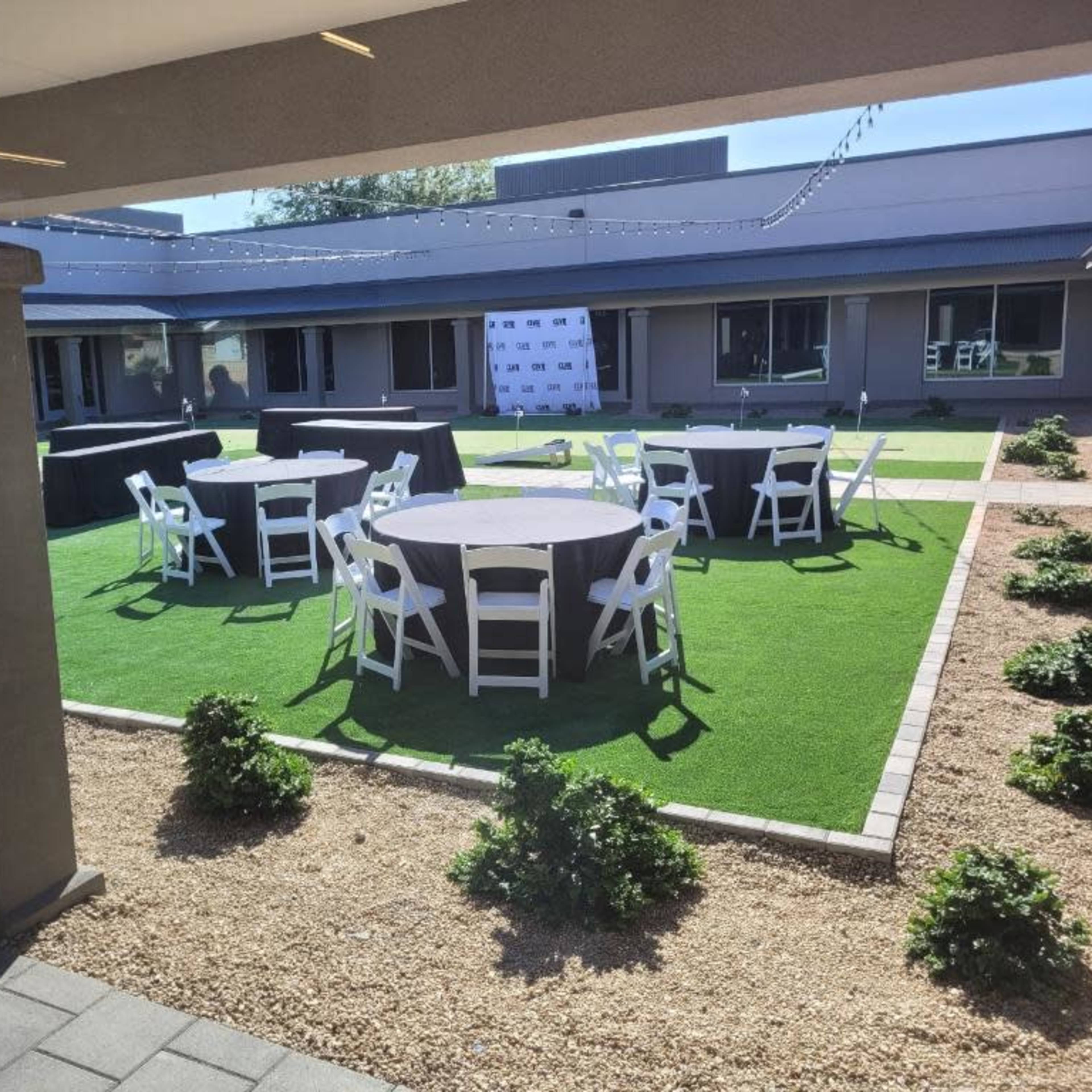 An outdoor event space with several round tables set up on grass and surrounded by hedges.