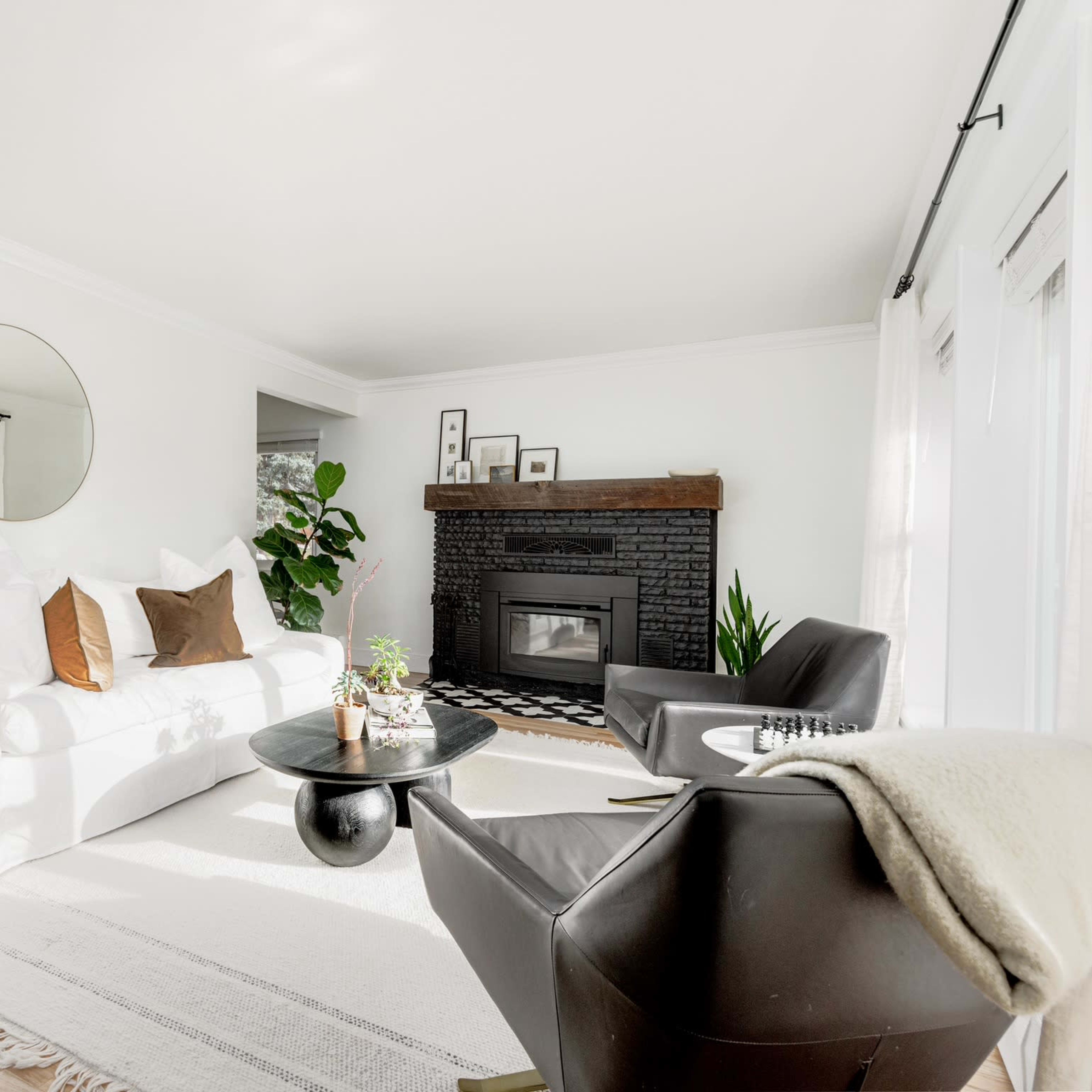 The living room features a white sofa, a black accent chair, a round coffee table, and a fireplace with a dark mantel, all set against light walls and wooden flooring.
