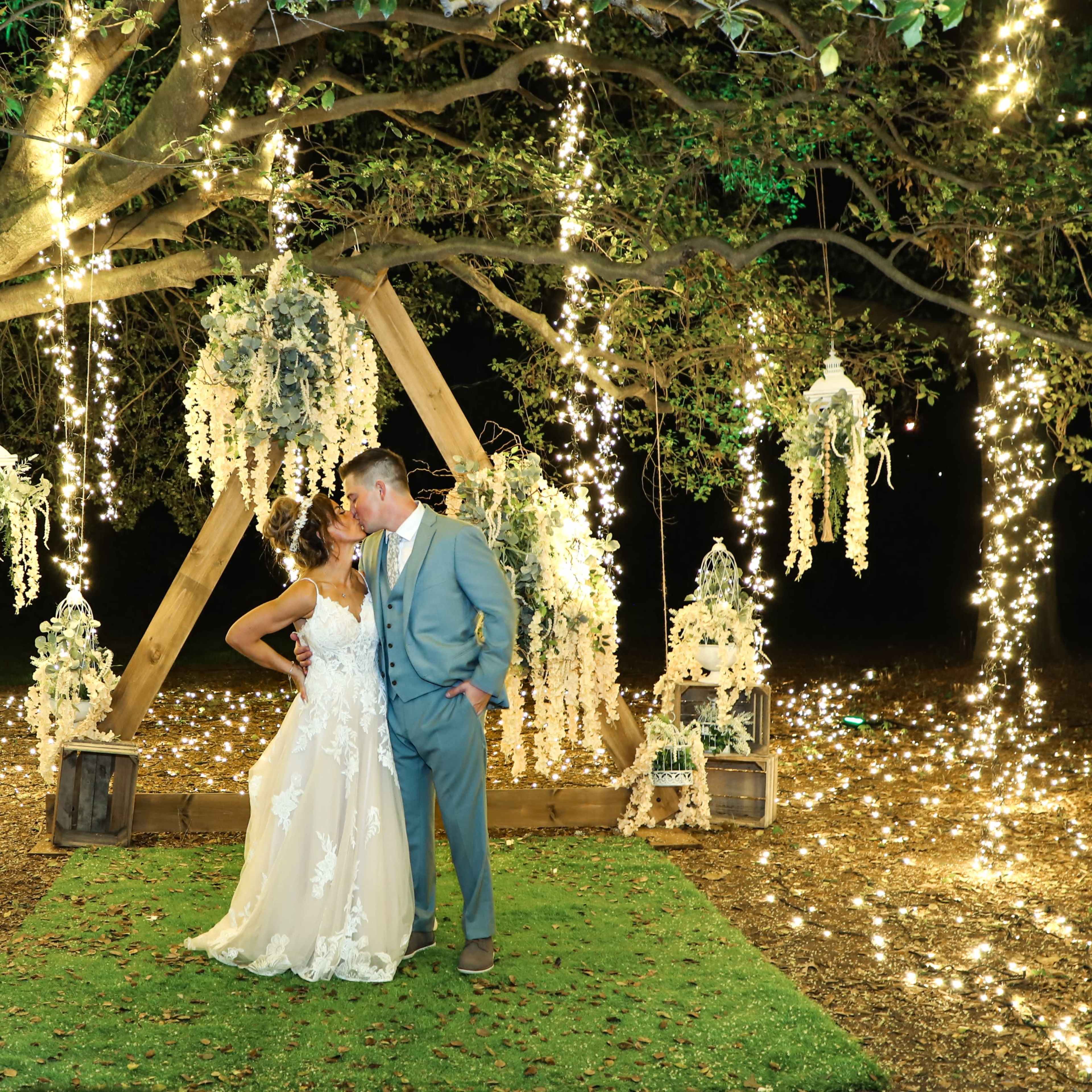 A couple in formal attire stands closely together under a decorated tree draped with lights and floral arrangements.
