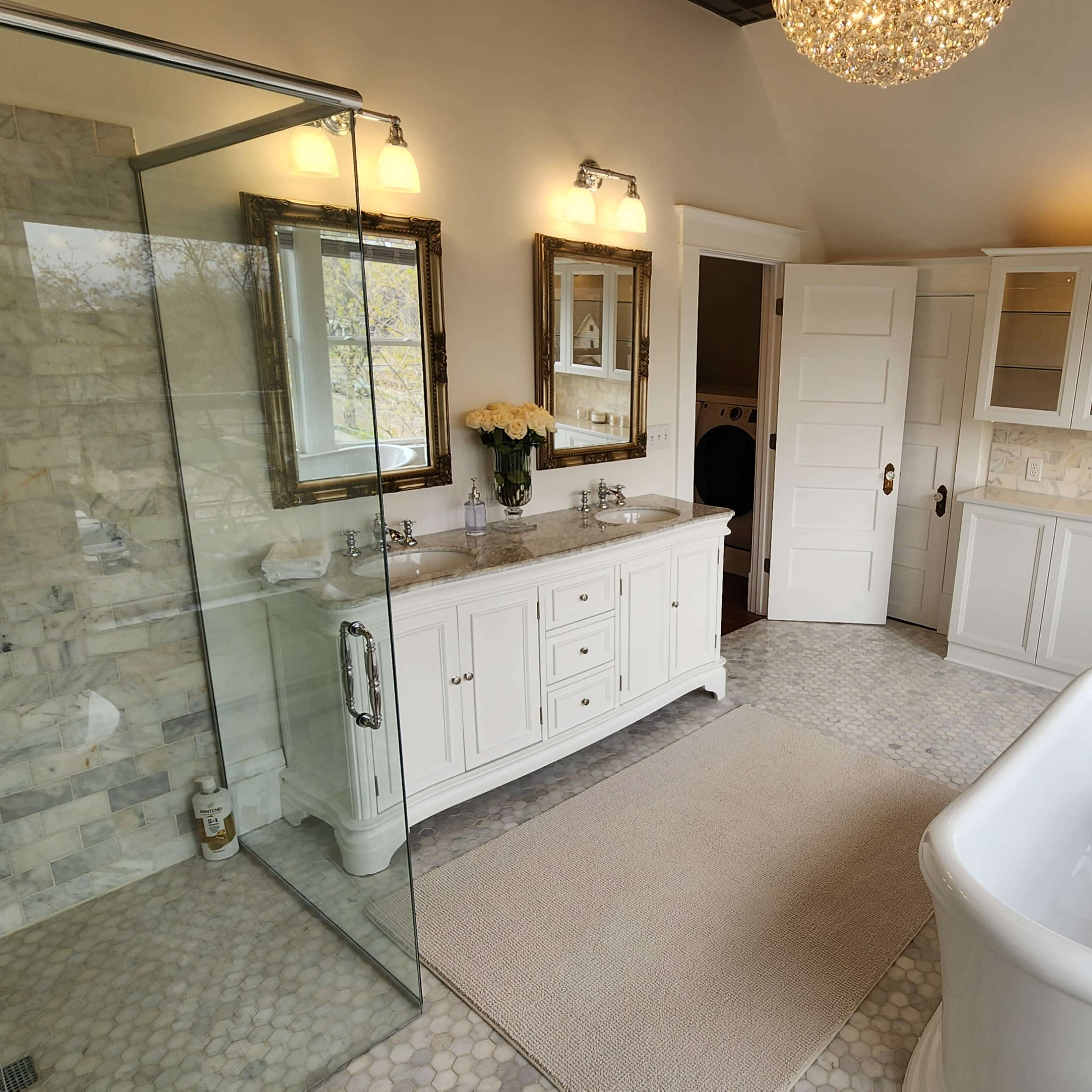 A bright, modern bathroom featuring a glass shower, a freestanding bathtub, double sinks in a white vanity, and a spacious layout with marble and stone finishes.