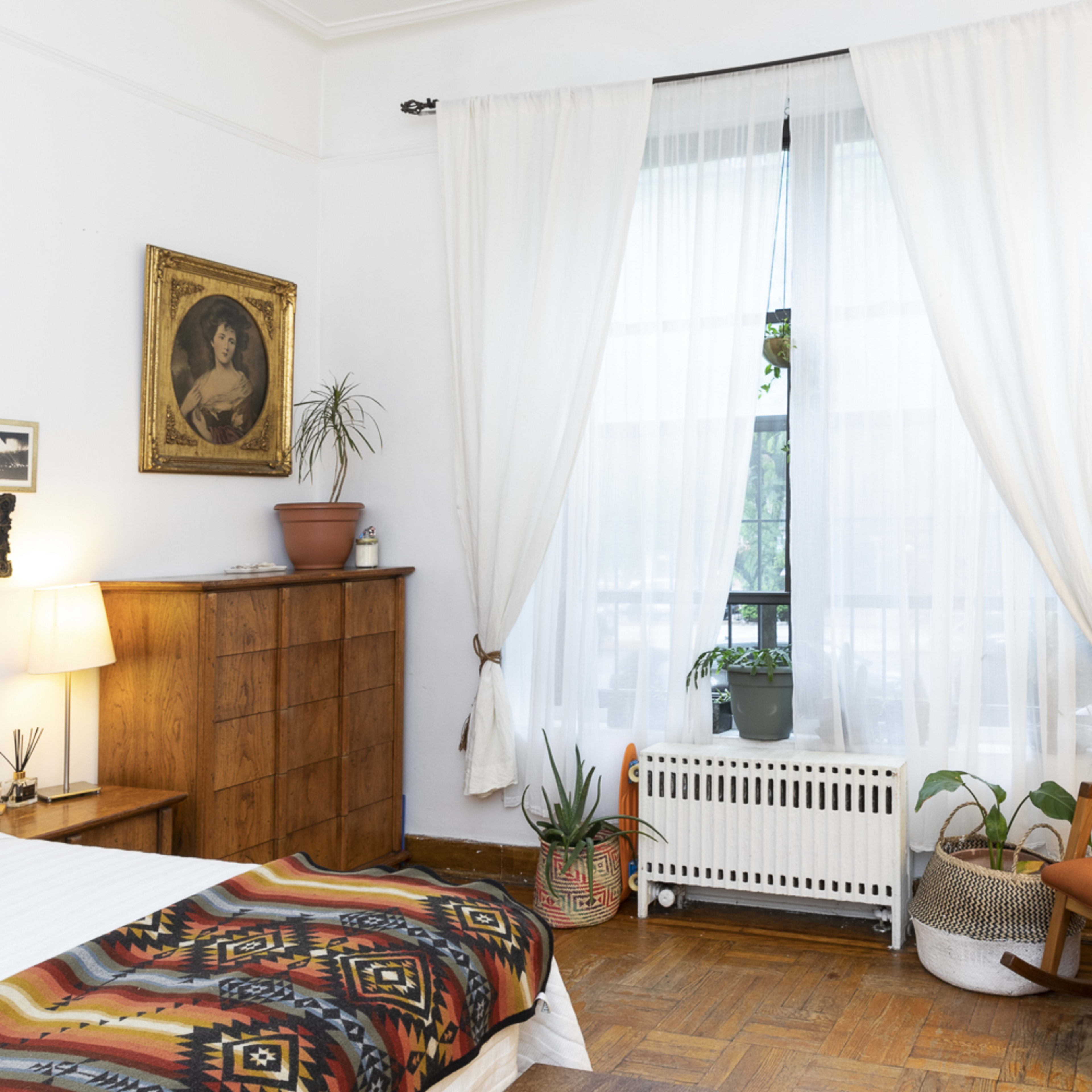 A cozy bedroom featuring a bed, a wooden dresser, potted plants, a rocking chair, and large curtains covering a window.