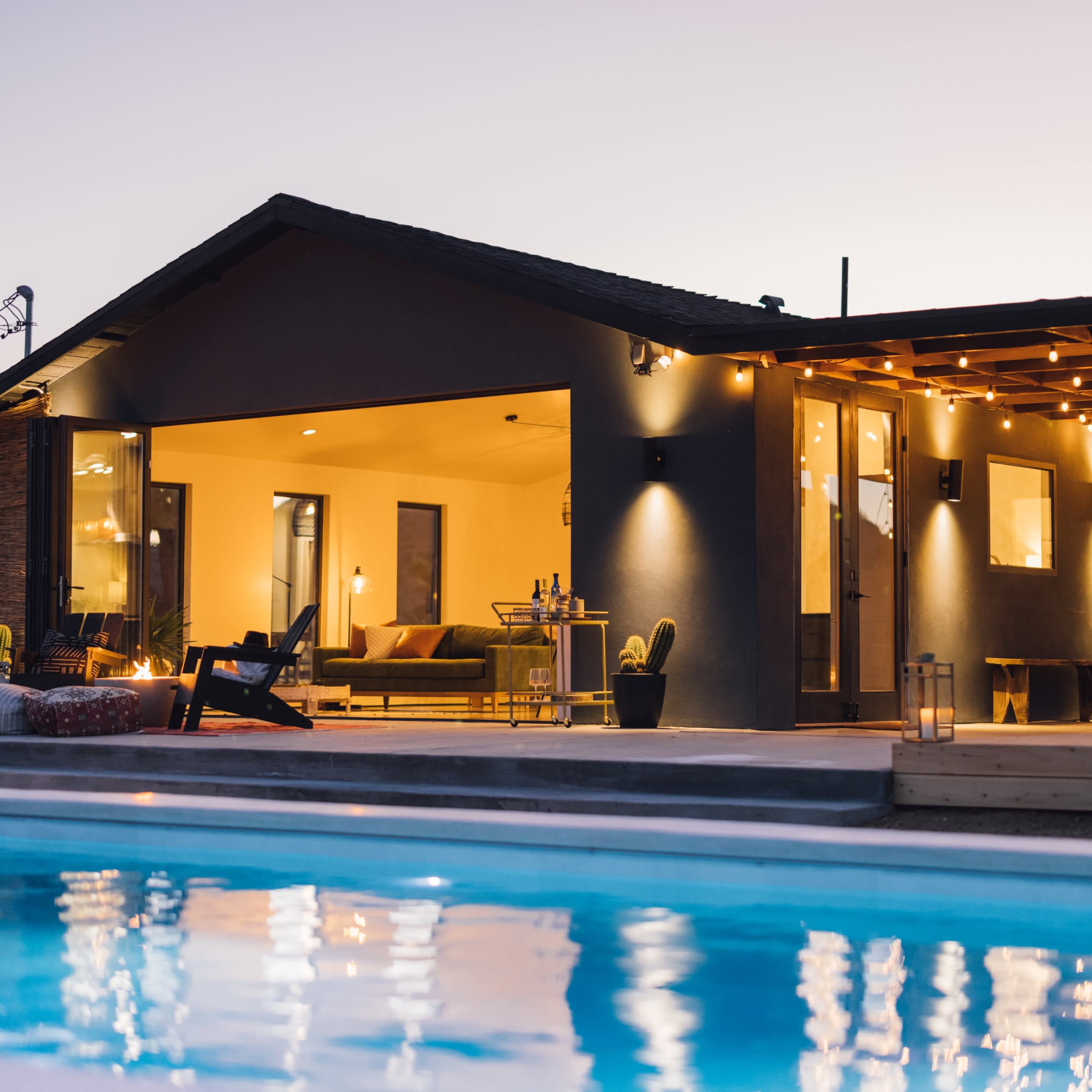 A modern house with large windows and outdoor seating overlooks a pool at dusk.