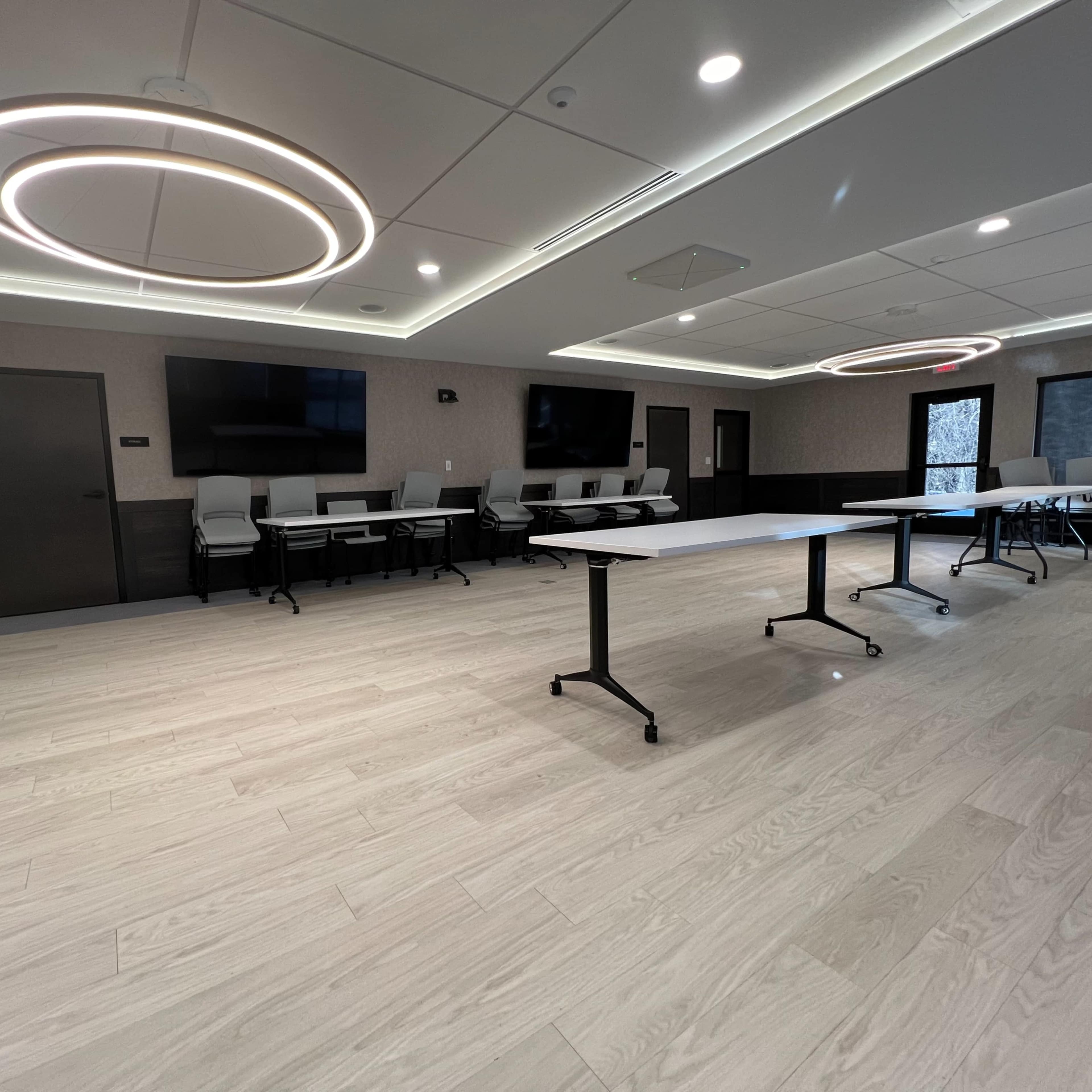 A modern meeting room with light wood flooring, two circular overhead lights, and tables arranged in the center.