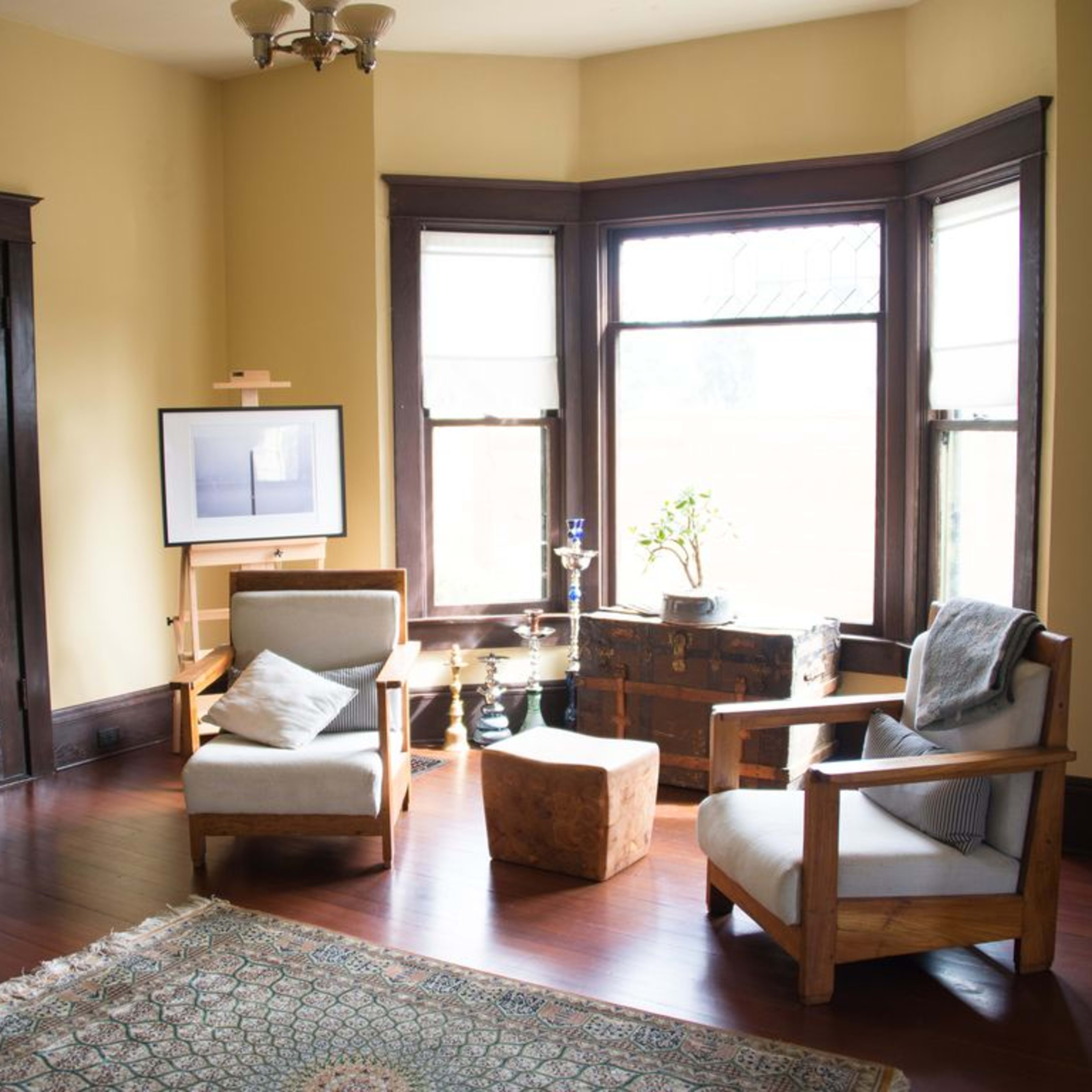 A cozy living room with two wooden chairs, a small ottoman, a flat-screen TV on an easel, and large windows allowing natural light.