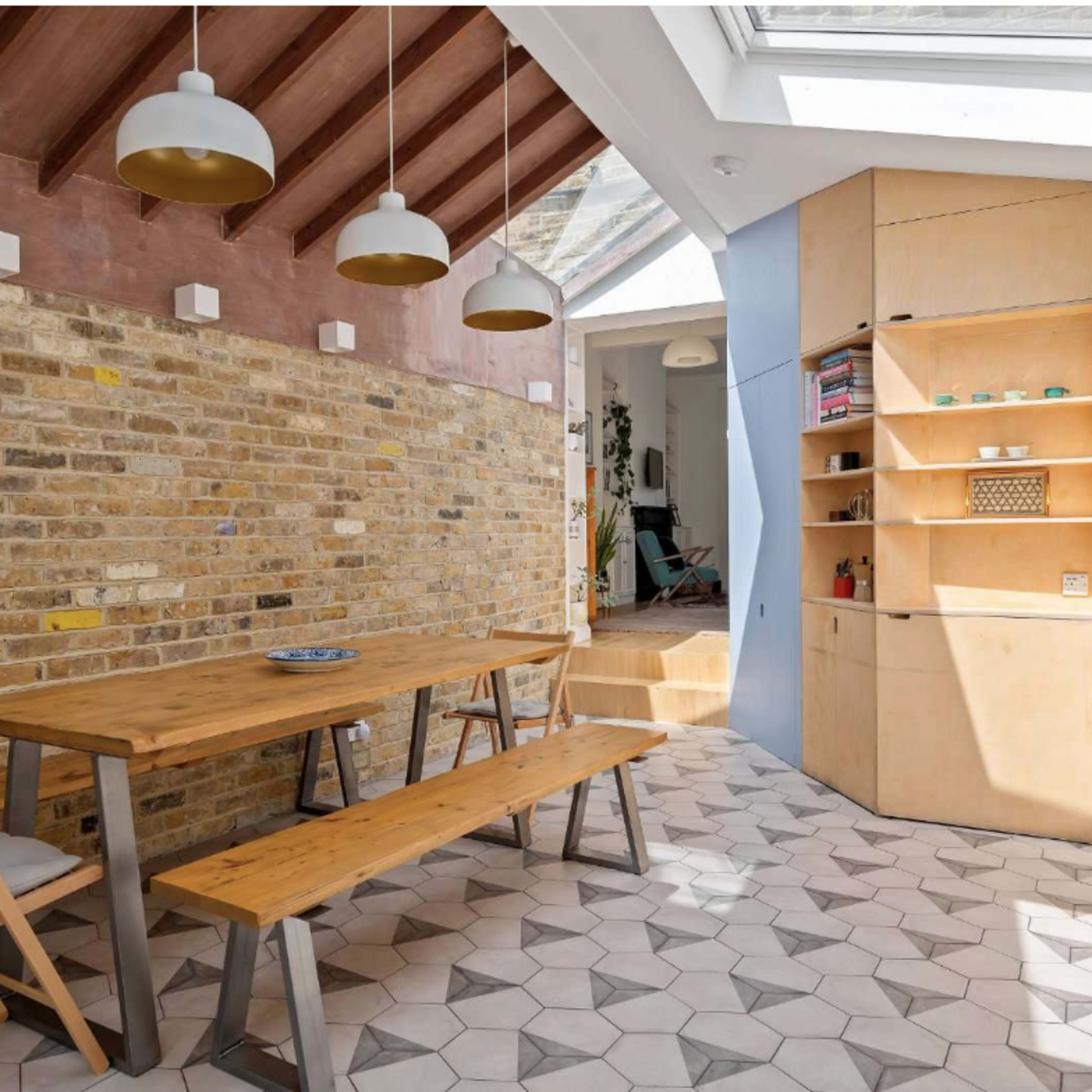 A modern interior dining area with a wooden table, benches, and a brick wall, complemented by geometric floor tiles and natural light coming through a skylight.
