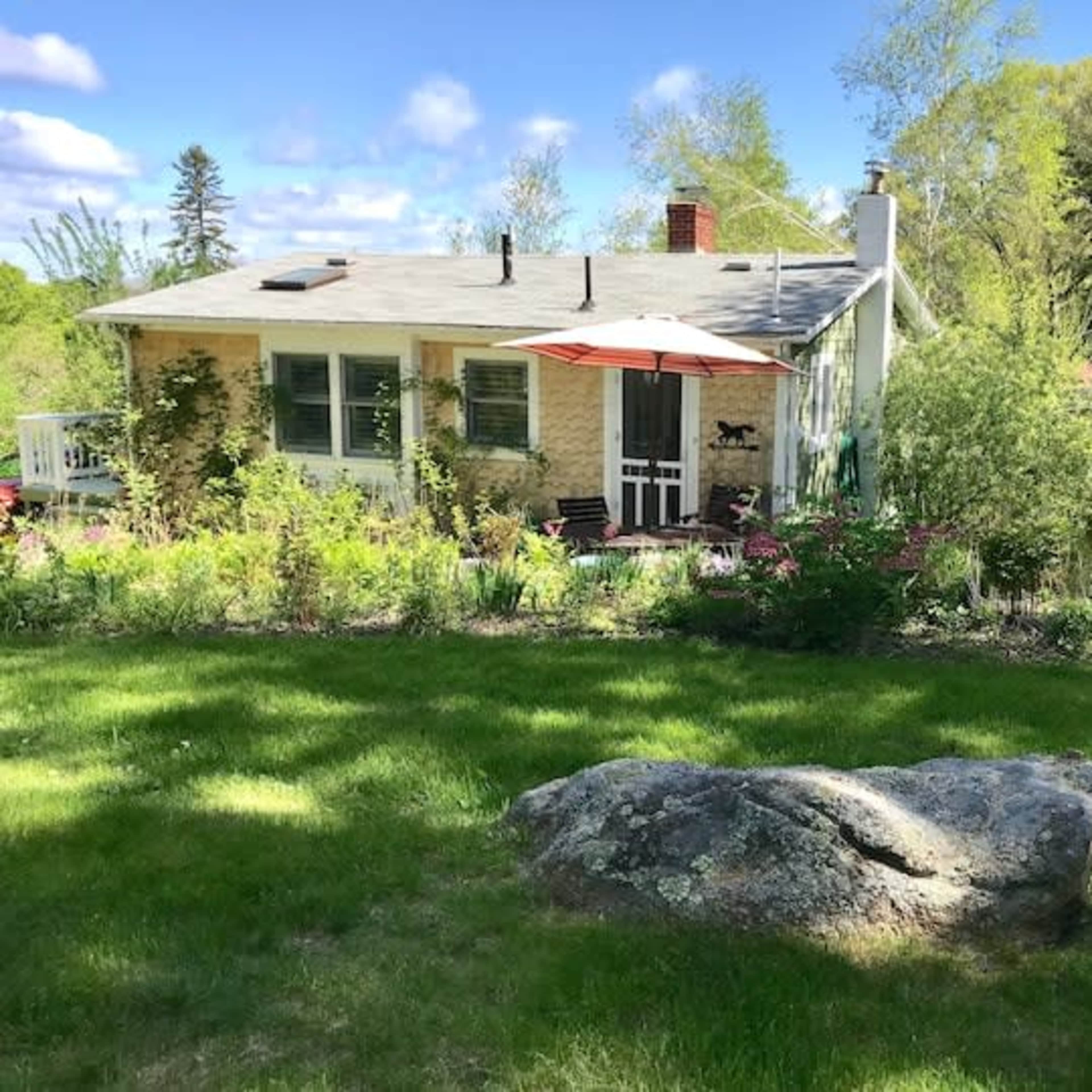 A single-story house with a gray roof and an orange umbrella stands in a garden surrounded by various plants and a large rock in the foreground.