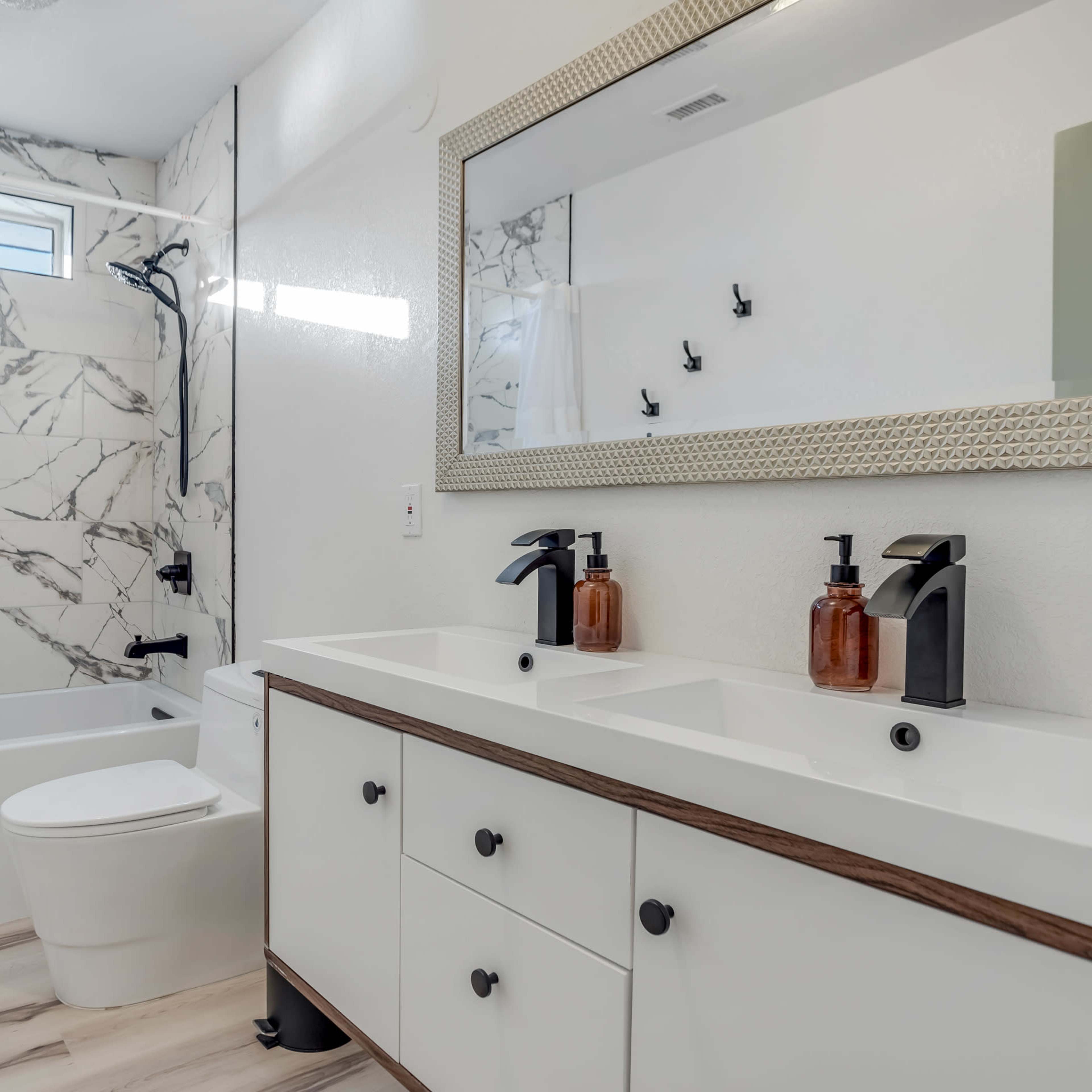 The bathroom features a marble-tiled wall, a modern double sink vanity with dark fixtures, a white bathtub, and a shower area with a glass partition.