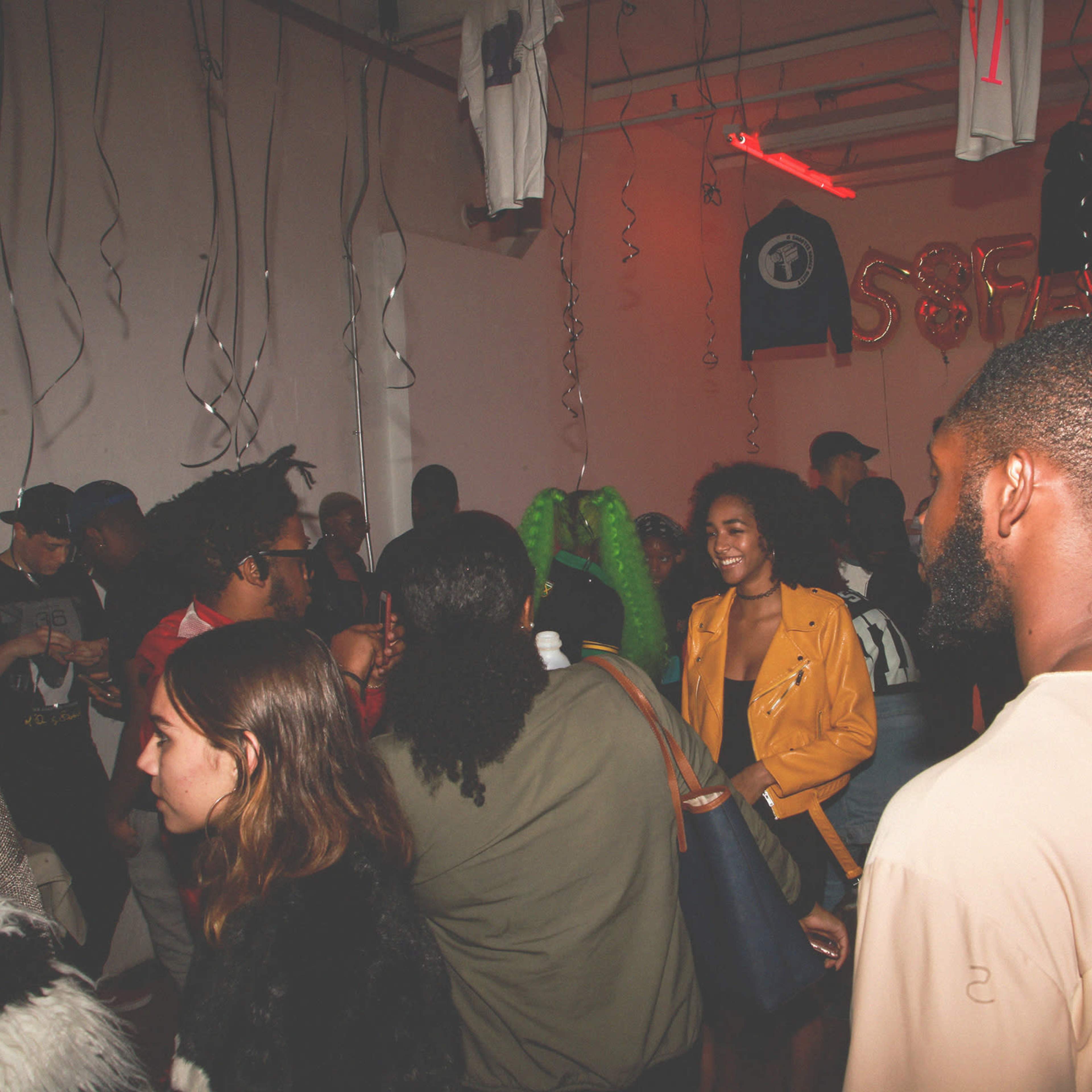 A diverse group of people socialize in a lively room decorated with balloons and hanging shirts.