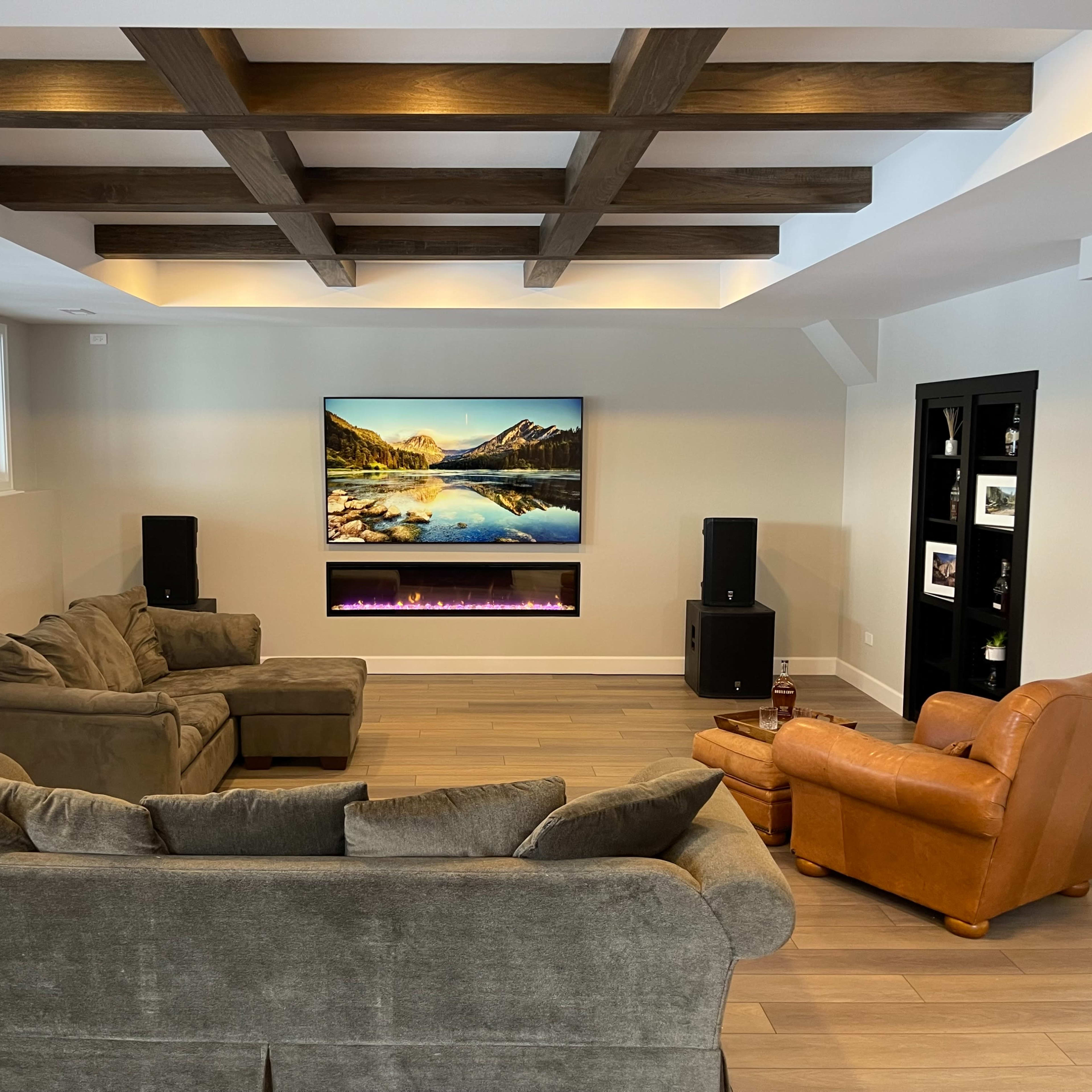 The room features a set of comfortable sofas arranged around a wall-mounted television with a scenic landscape display and an electric fireplace below it.
