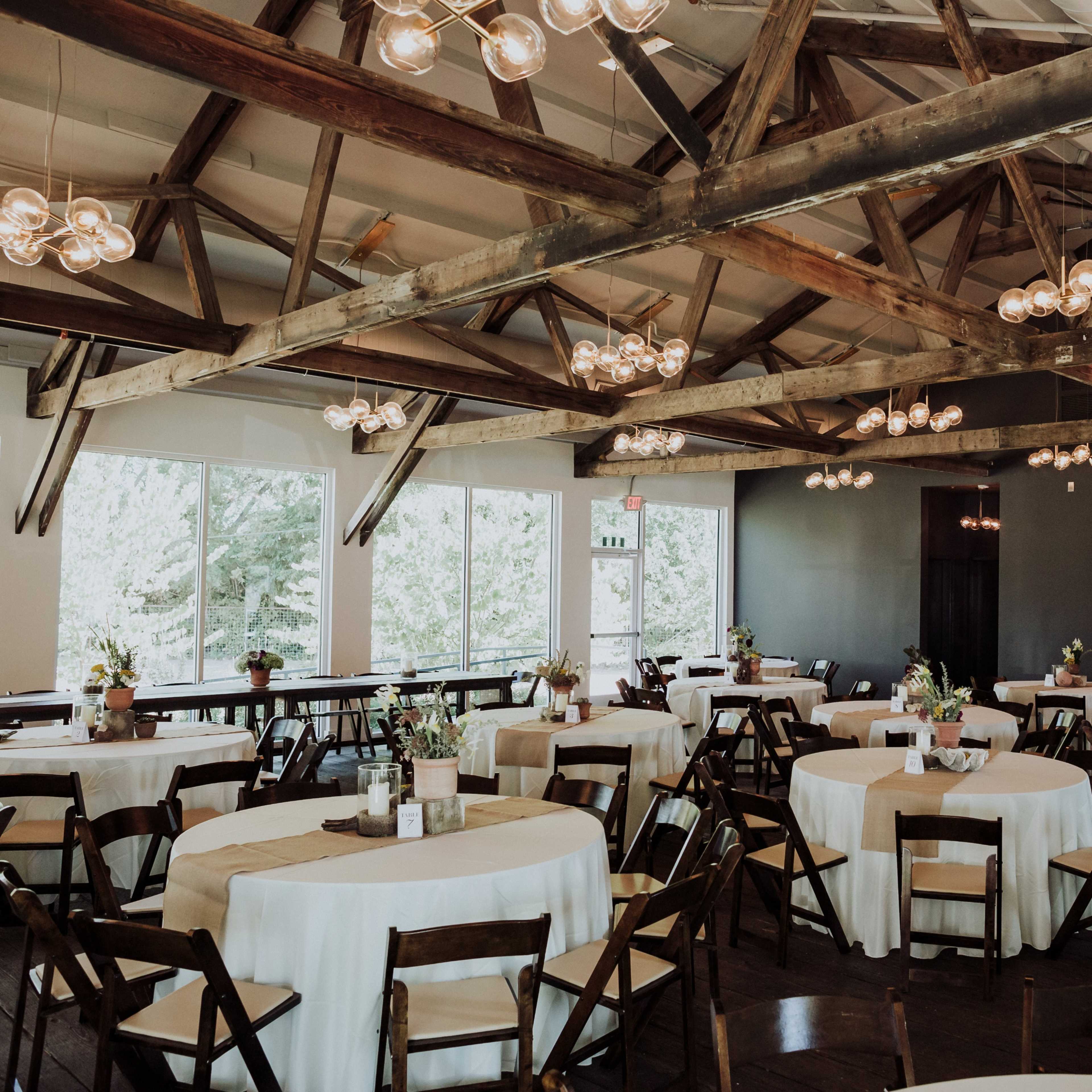 A spacious event venue with round tables set for dining, wooden beams overhead, and large windows allowing natural light to enter.