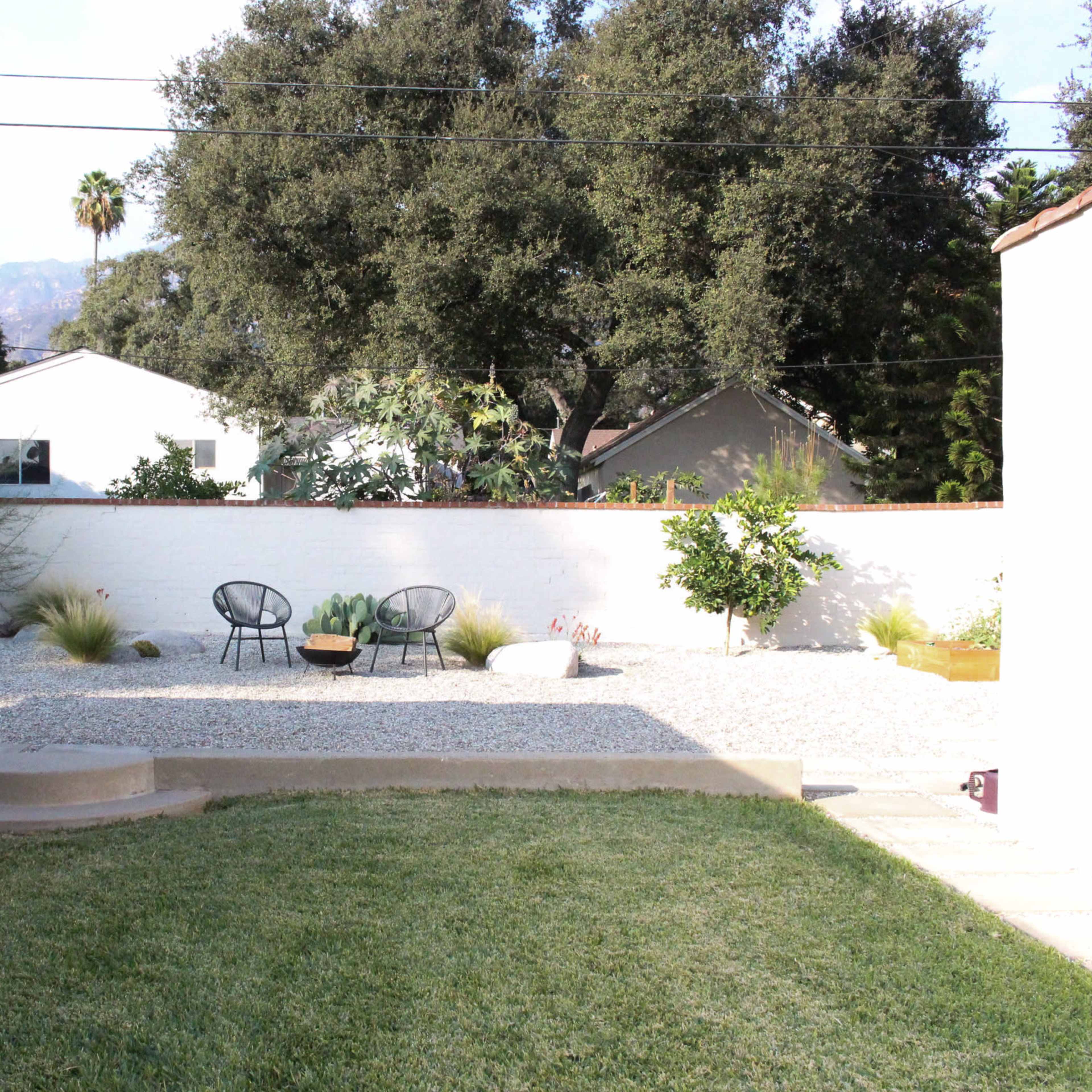 A backyard features a gravel landscape with two black chairs and a small tree in the background, surrounded by low vegetation and a white wall.