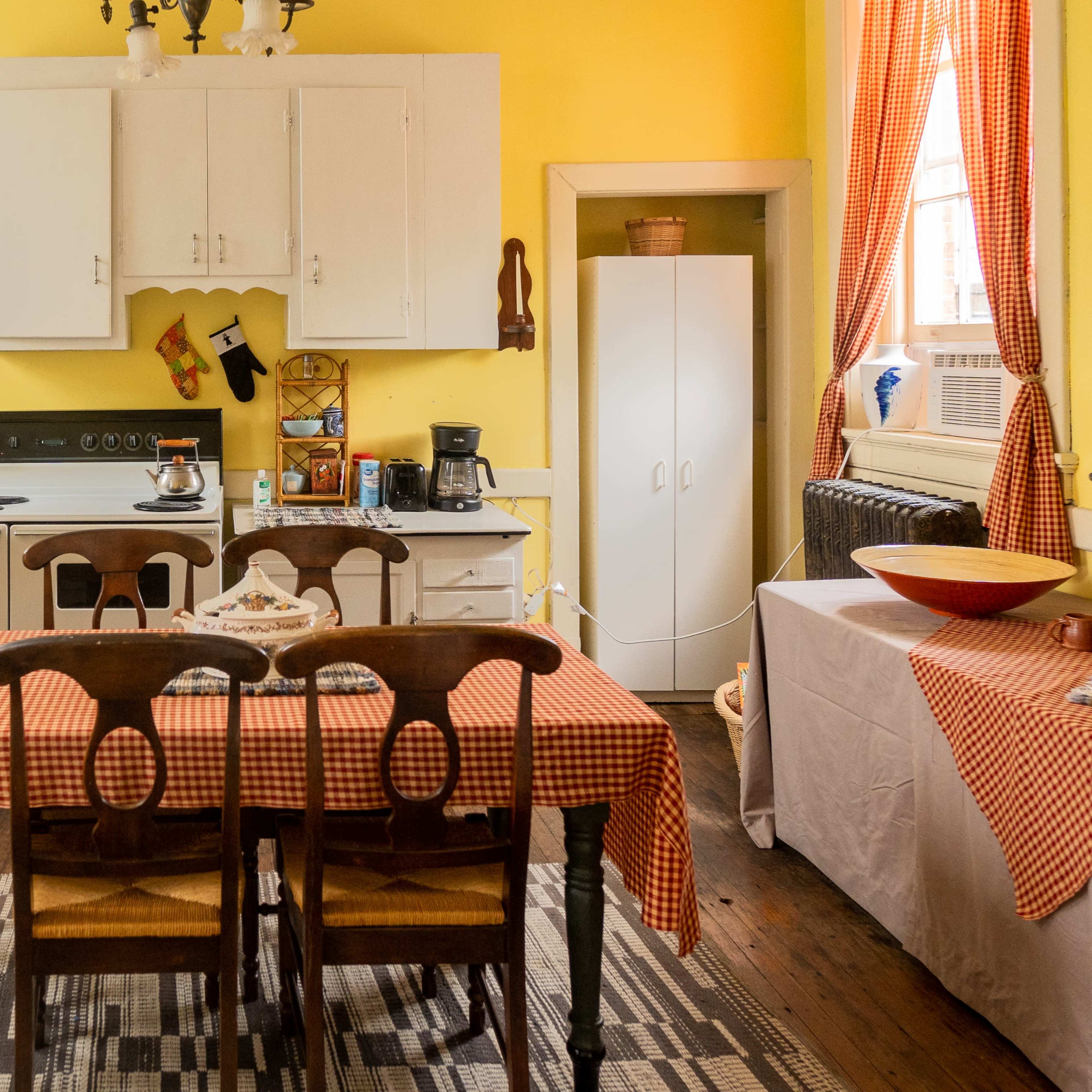 A kitchen with yellow walls, a central table covered in a checkered tablecloth, and white cabinets along with a stove and coffee maker.