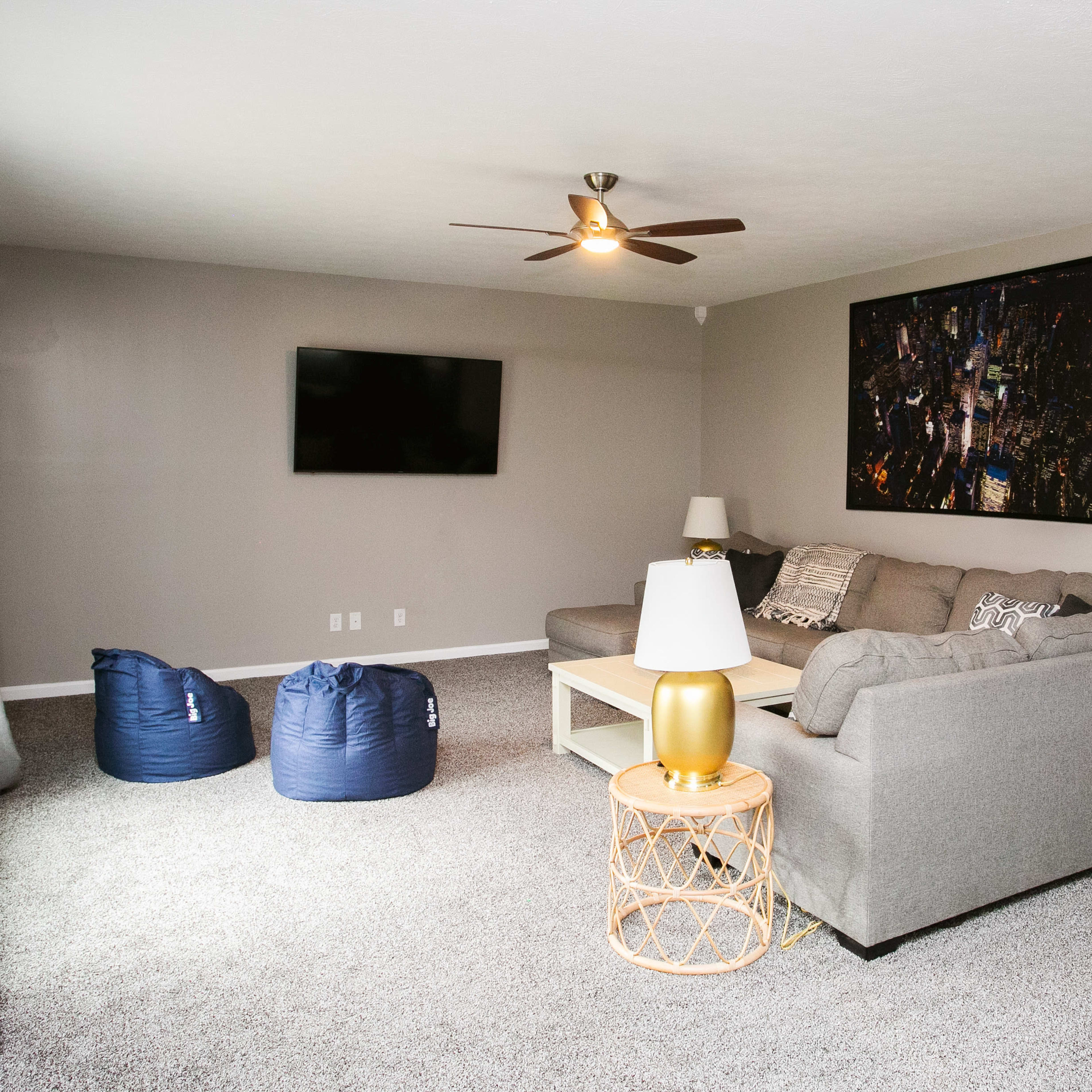 The living room features a gray sofa, a wall-mounted TV, a coffee table, two blue bean bags, and a lamp on a gold side table, all set against neutral walls and a carpeted floor.