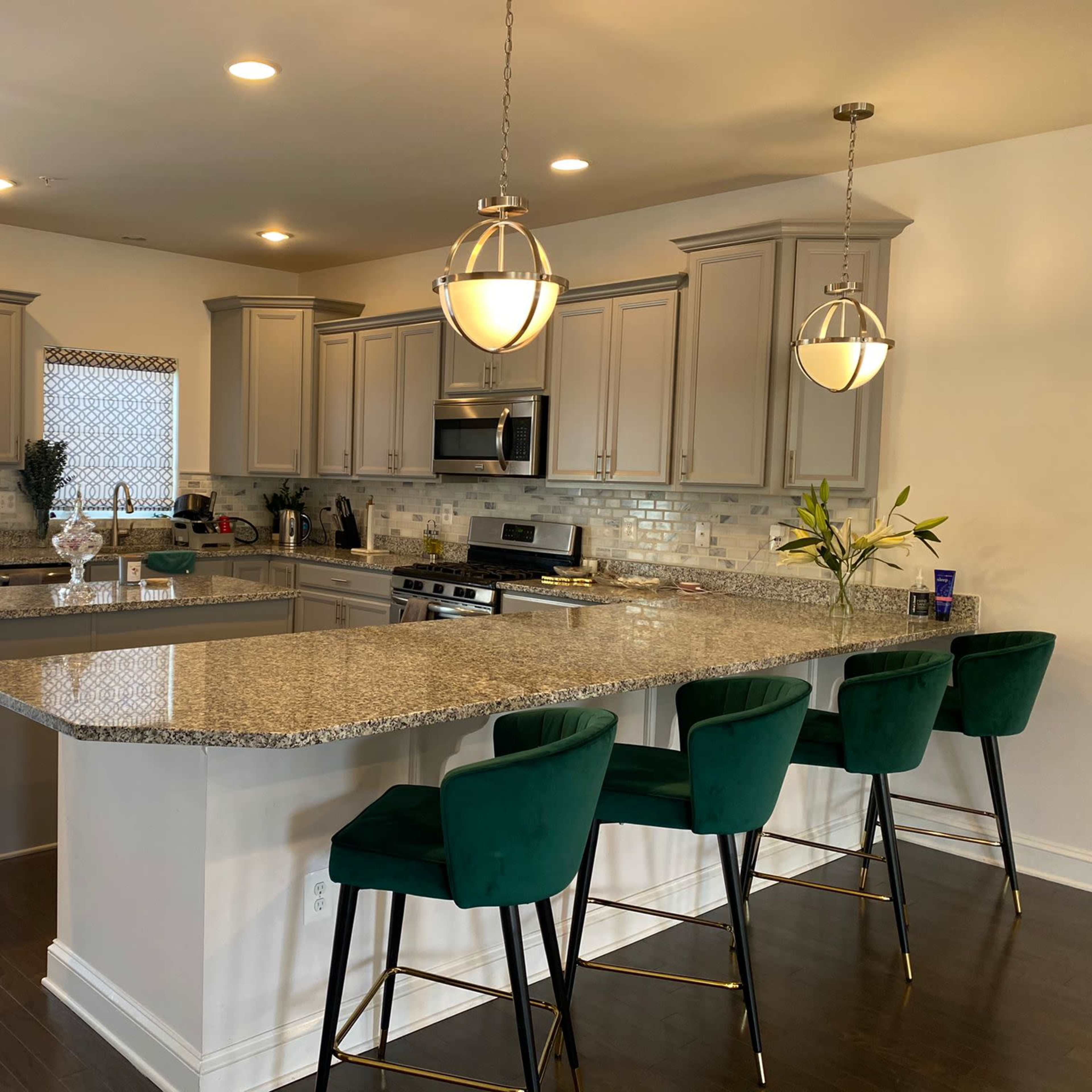 A modern kitchen features gray cabinetry, granite countertops, stainless steel appliances, and four green bar stools at a large island.