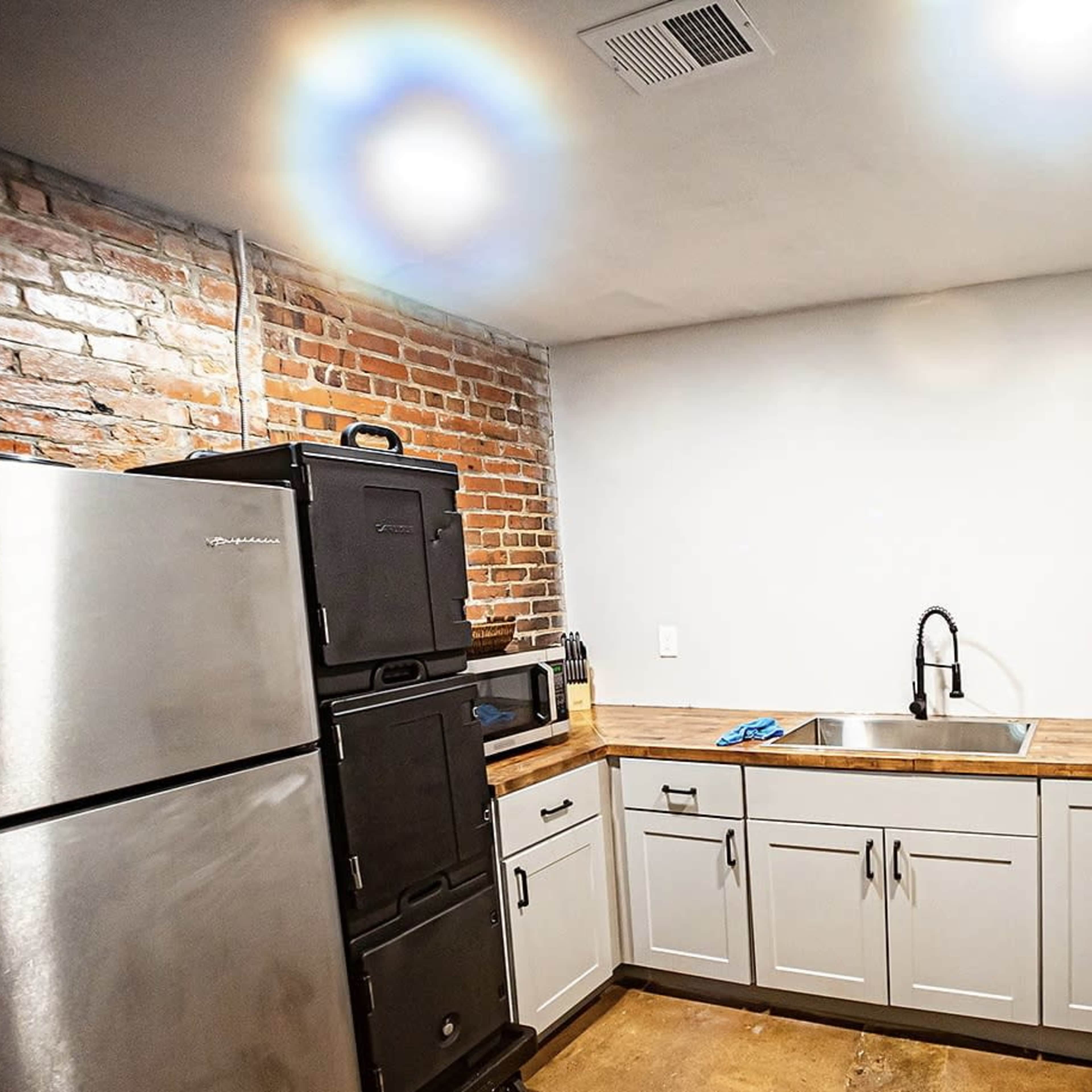 A kitchen area with exposed brick walls, a stainless steel refrigerator, and a wooden countertop with a sink and gray cabinets below.