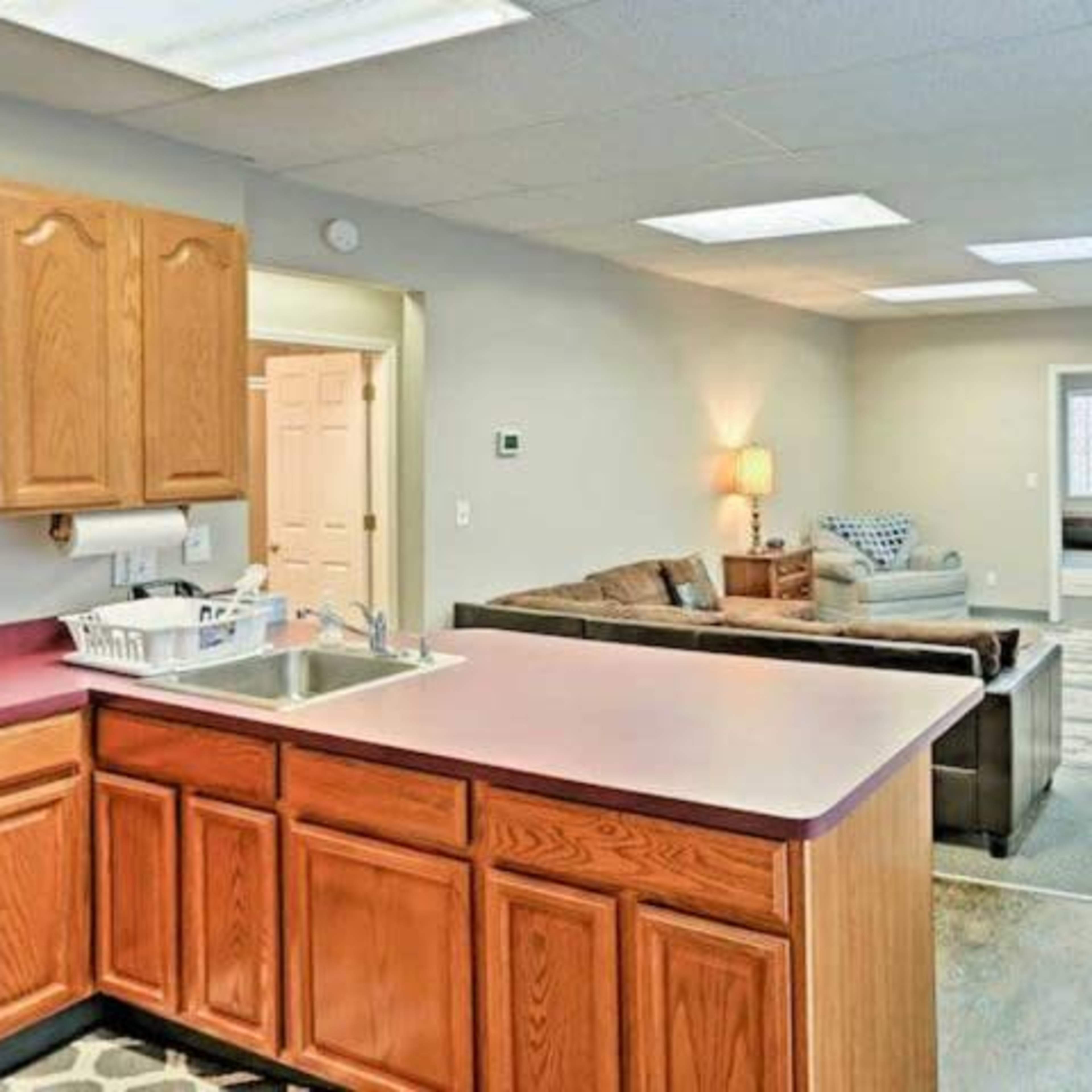 A kitchen area with wooden cabinets adjoins a living space featuring a couch, a television, and a light-colored carpet.