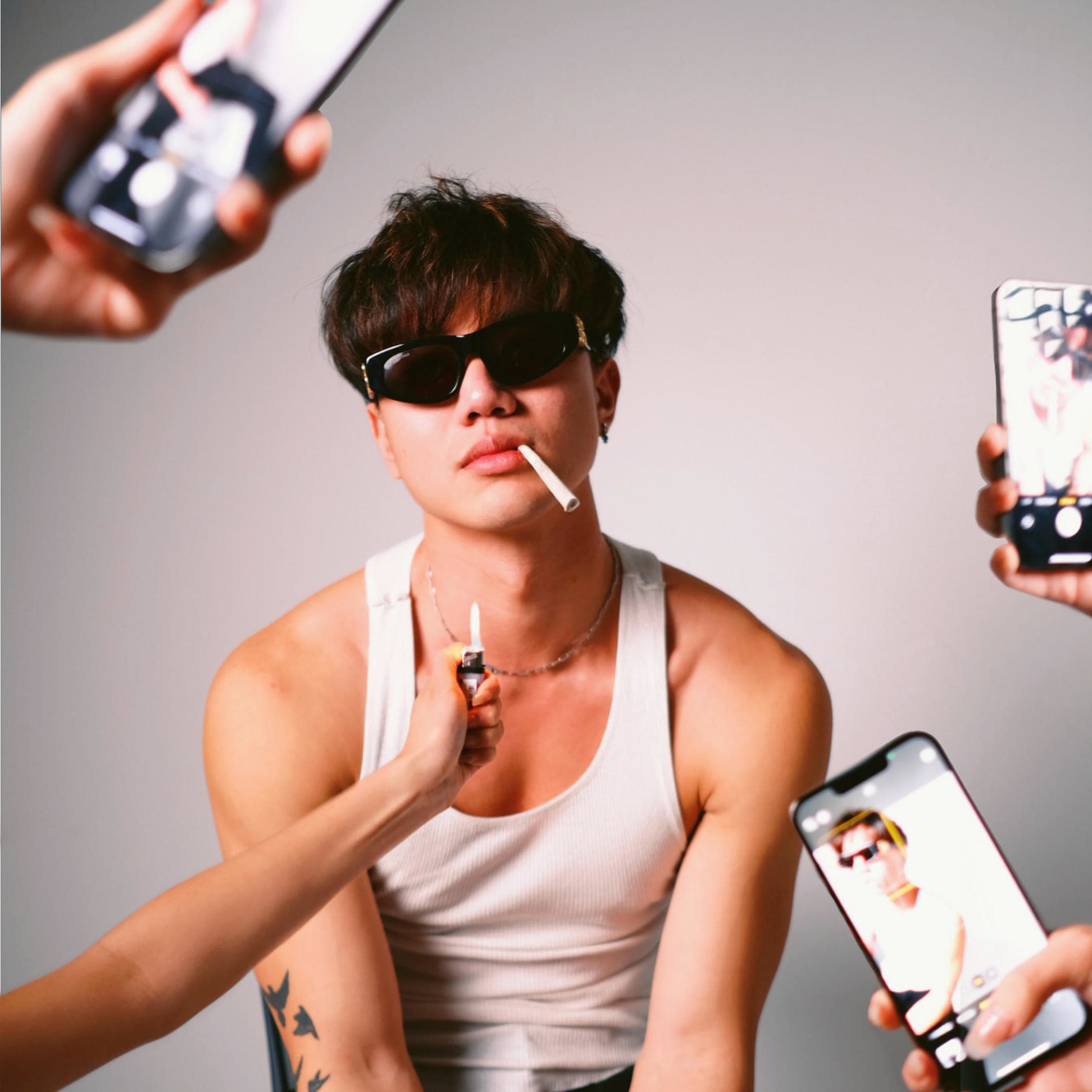 A young man wearing sunglasses and a tank top poses with a cigarette, while several hands hold smartphones recording him.