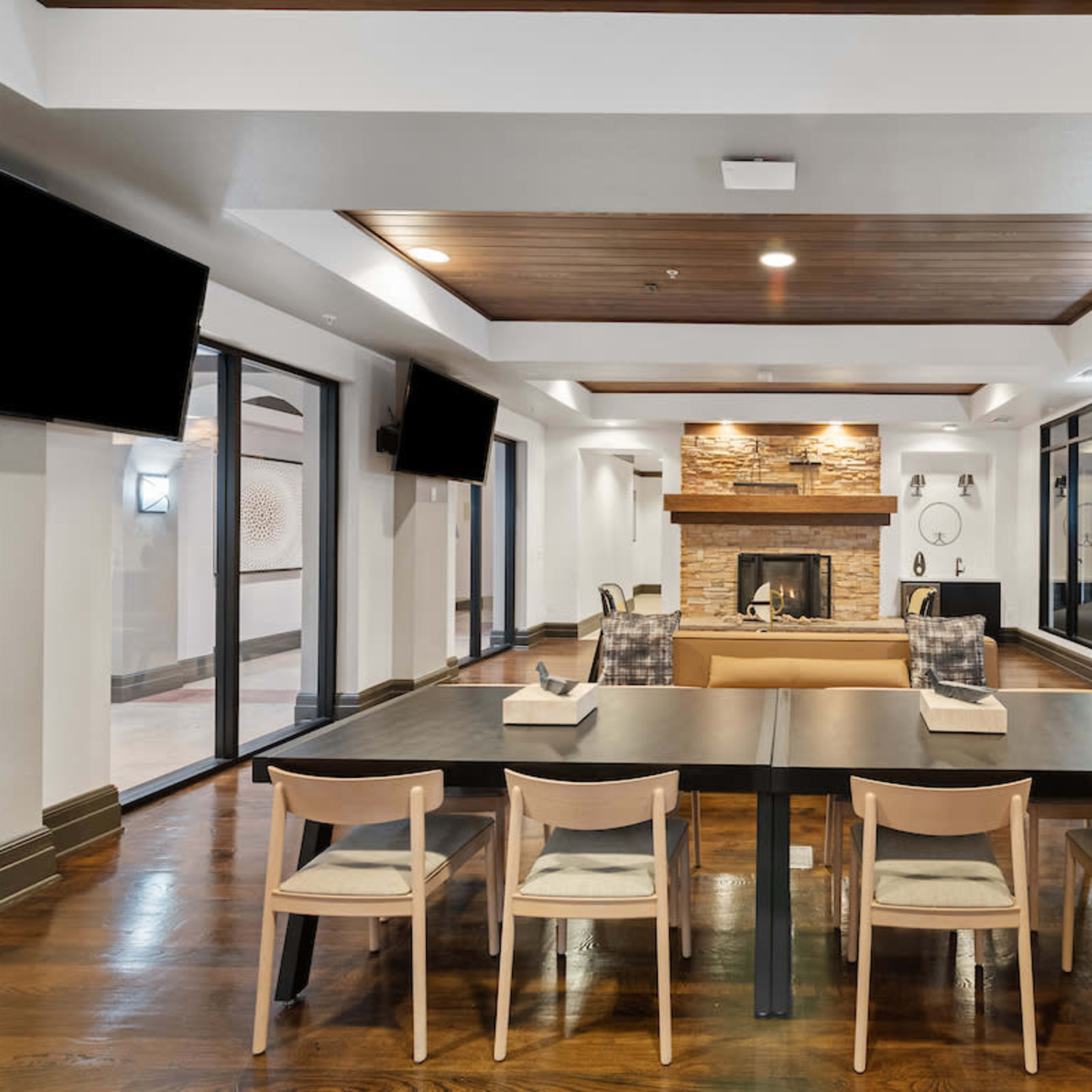 A modern communal room features a central dining table with chairs, a fireplace with a stone surround, and large windows providing natural light.