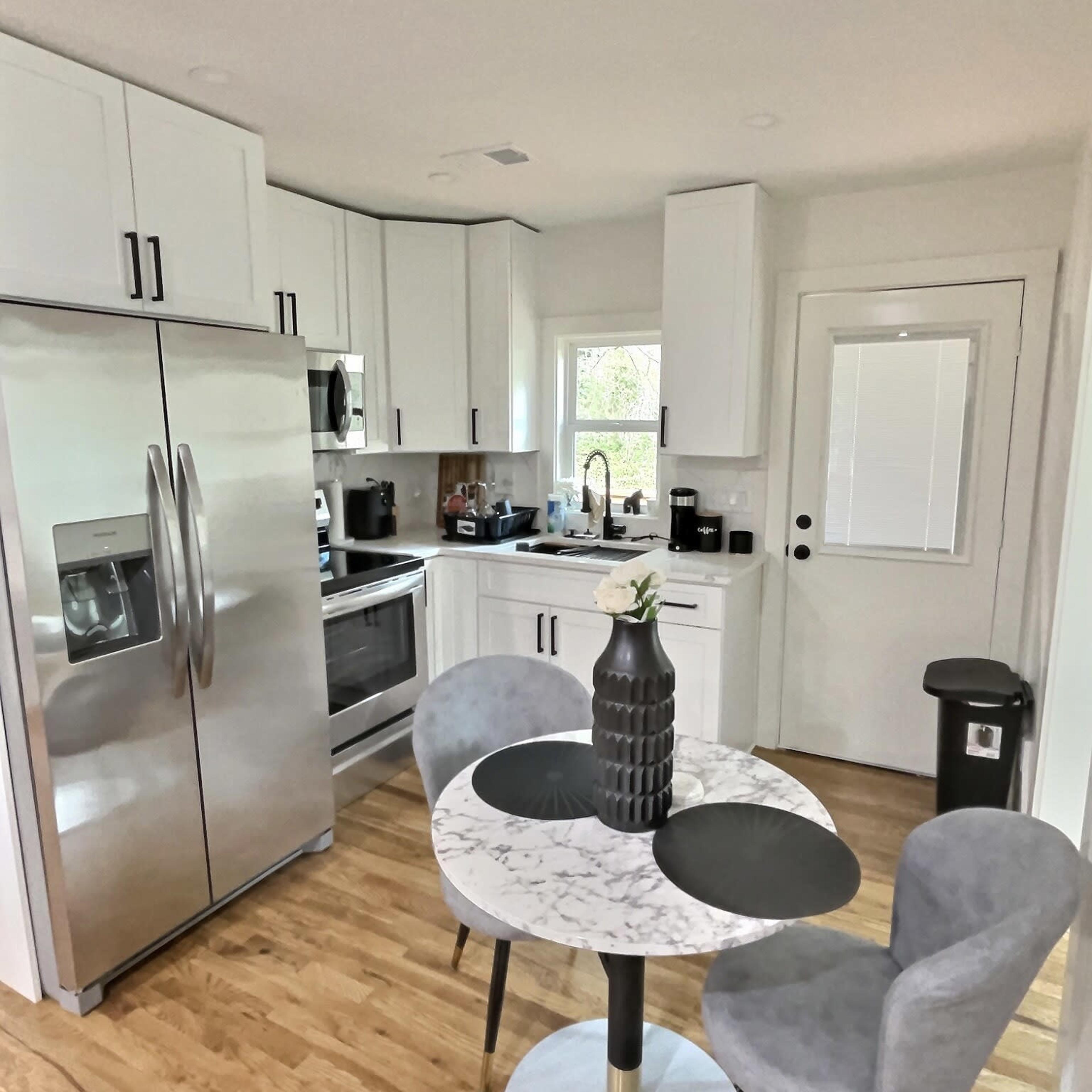A modern kitchen featuring stainless steel appliances, a round marble dining table with two chairs, and a window providing natural light.
