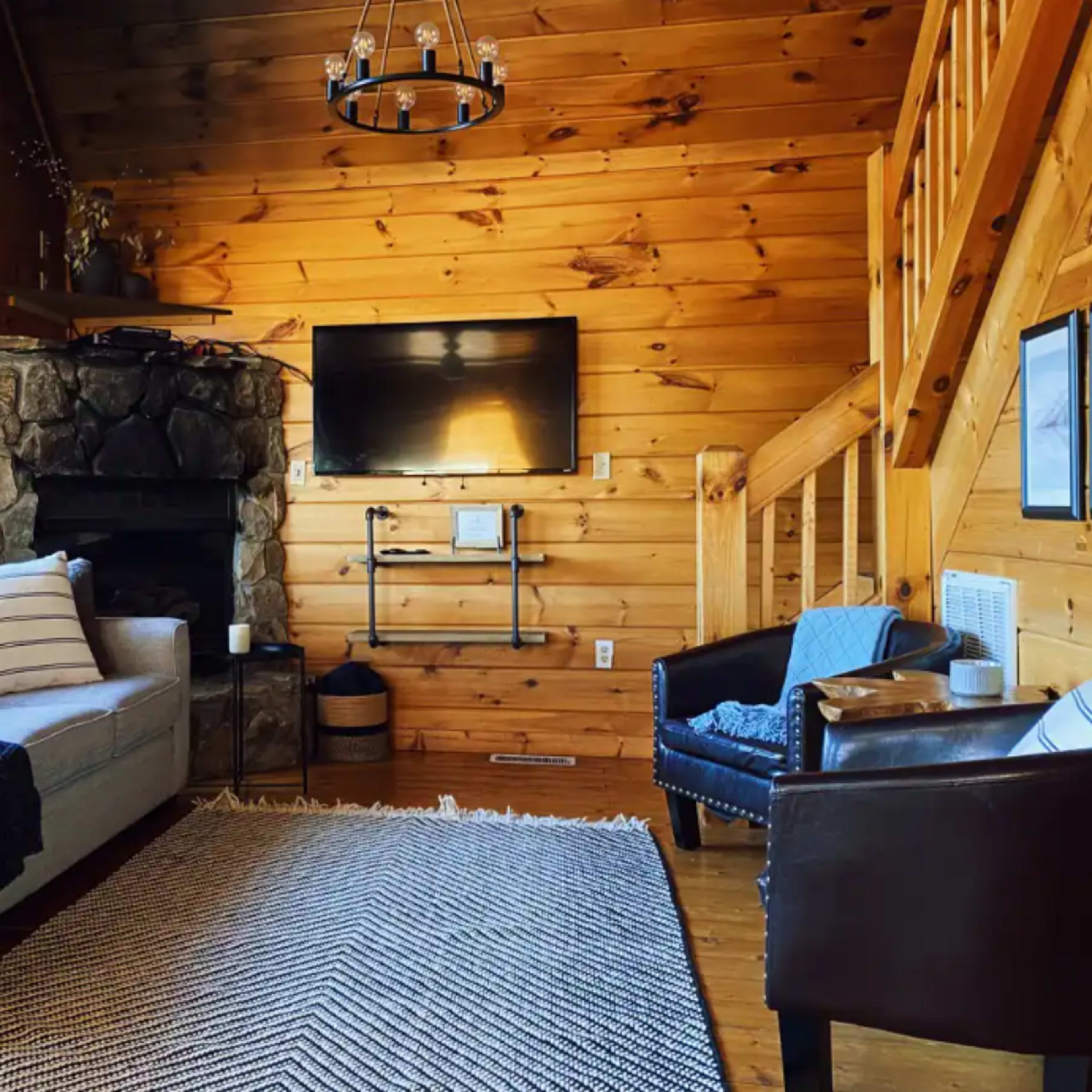 The living room features a gray sofa, a stone fireplace, a mounted television, wooden walls, and a staircase.