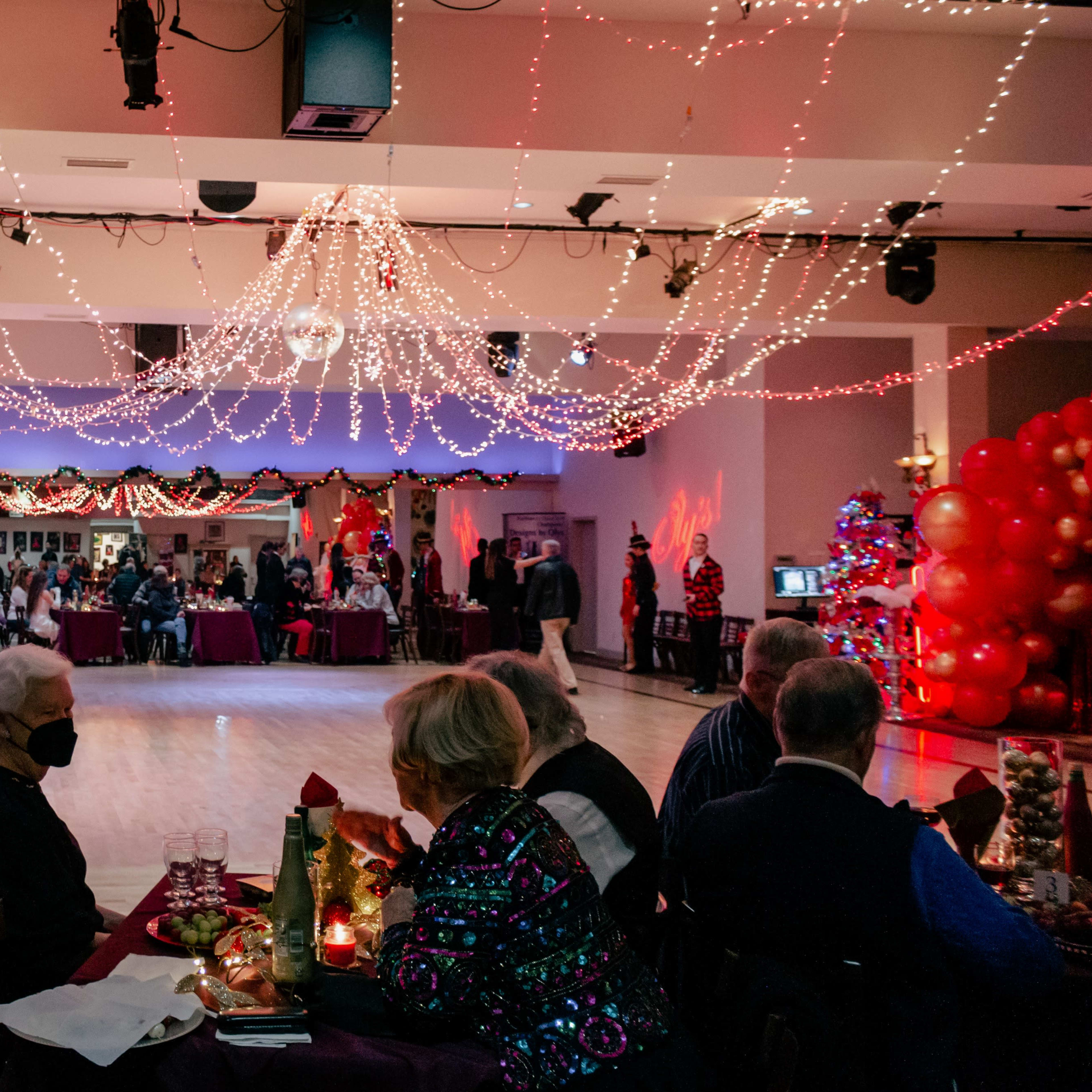 A festive dining event is taking place in a decorated hall with twinkling lights and tables filled with guests.