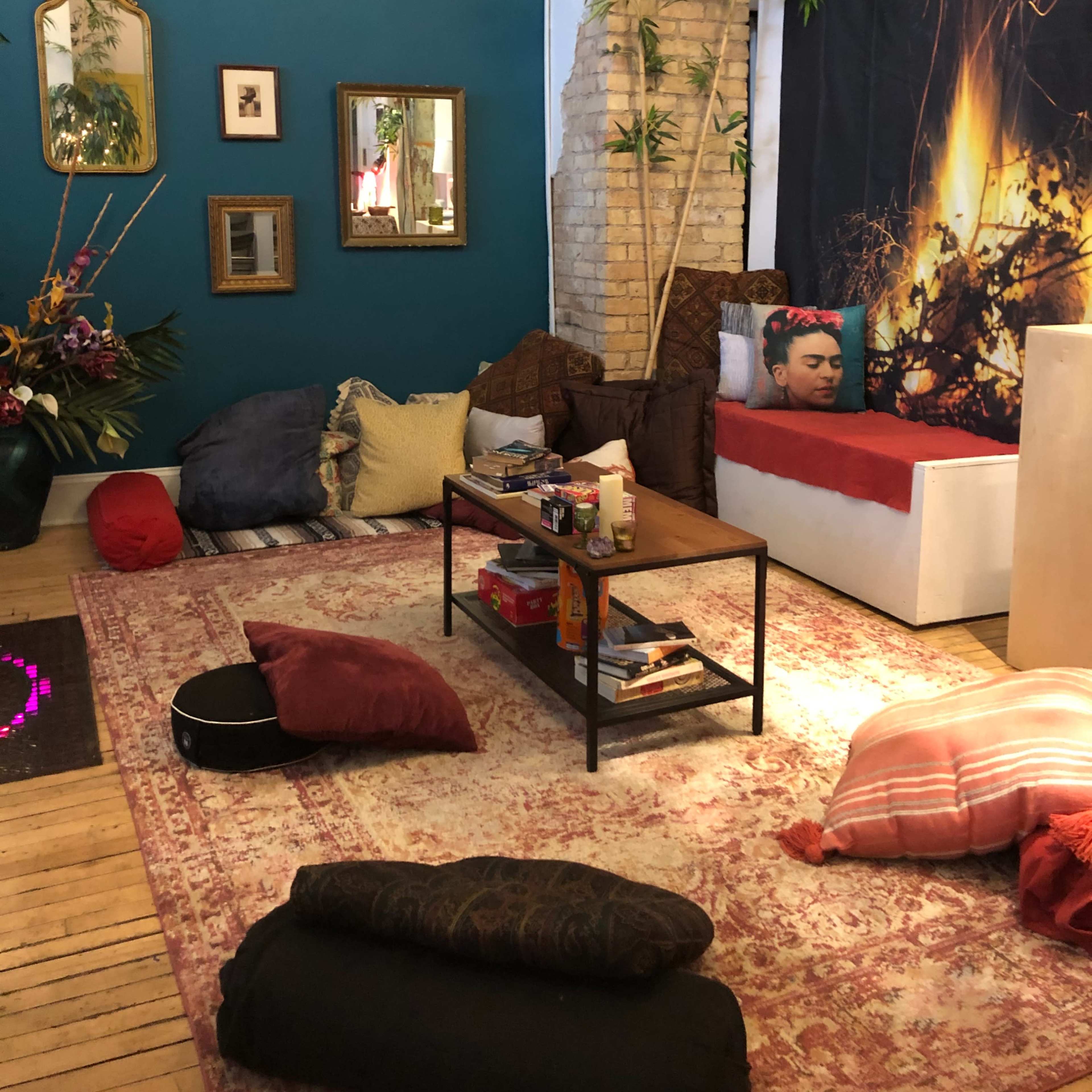 The room features a mix of seating options including pillows and a low table, with a red bedspread and a wall adorned with plants and artwork.