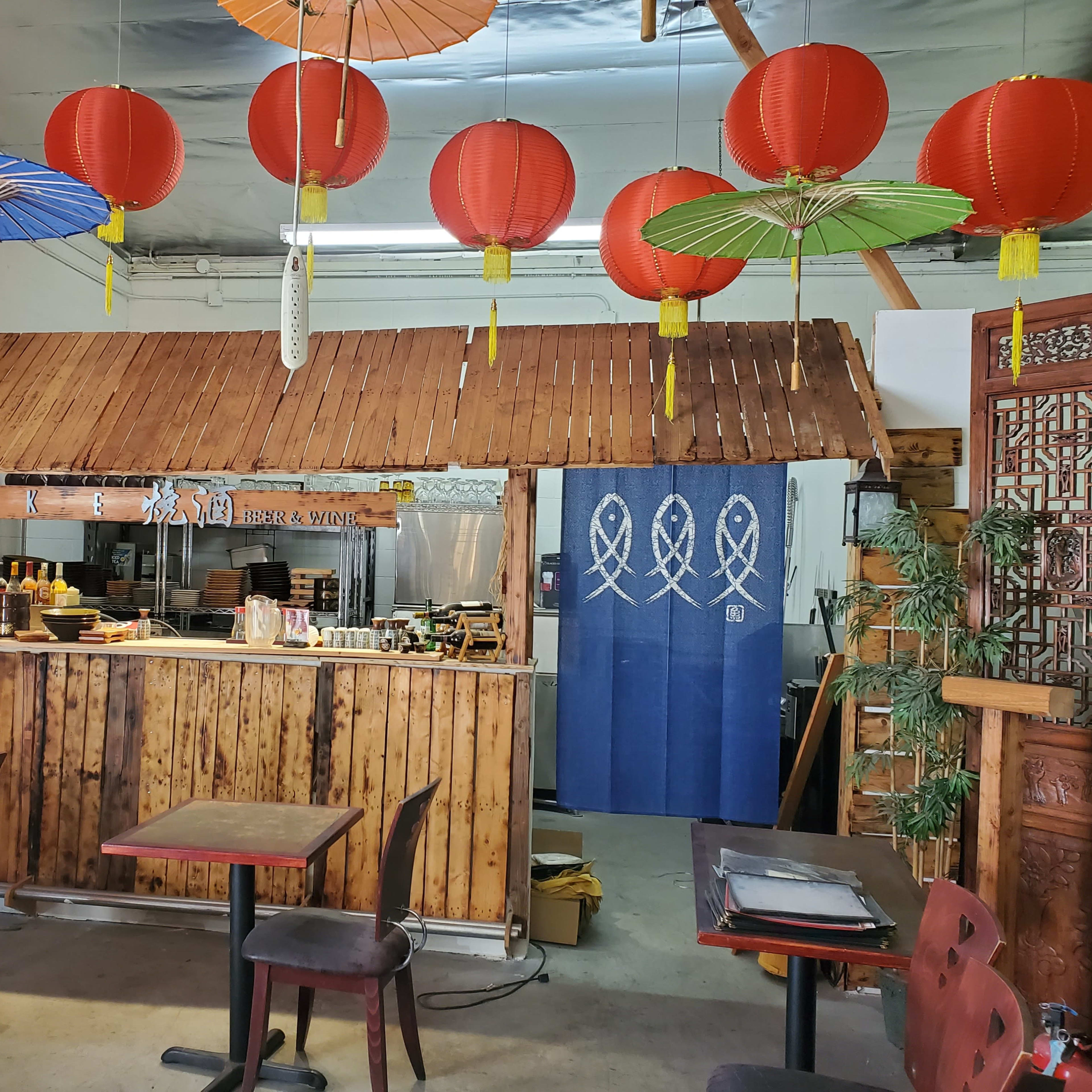 A wooden counter with hanging red lanterns and colorful paper umbrellas is seen in an Asian-themed restaurant.