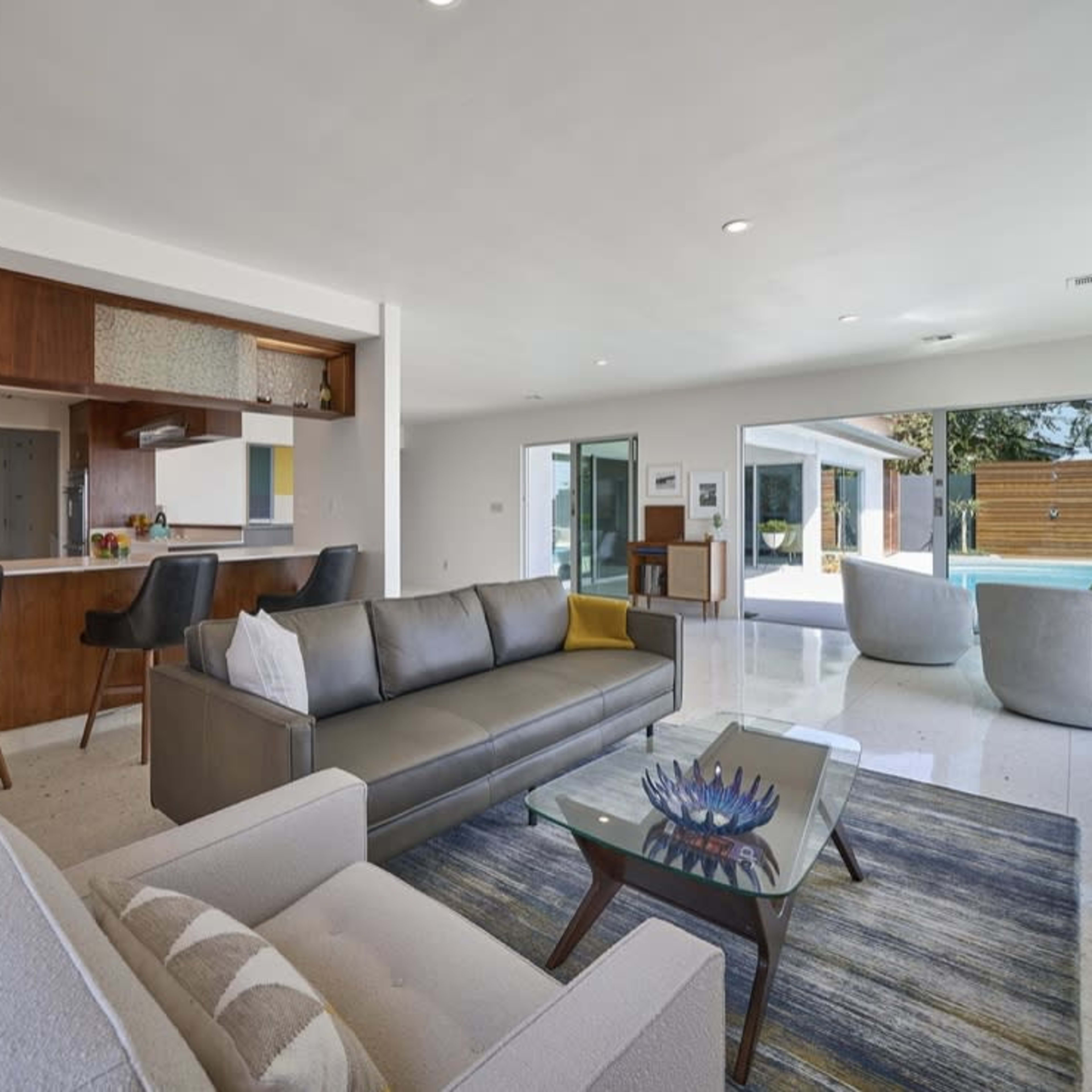 A modern living room with a gray sofa, an armchair, a glass coffee table, and an open kitchen area featuring dark wood cabinetry and bar stools.