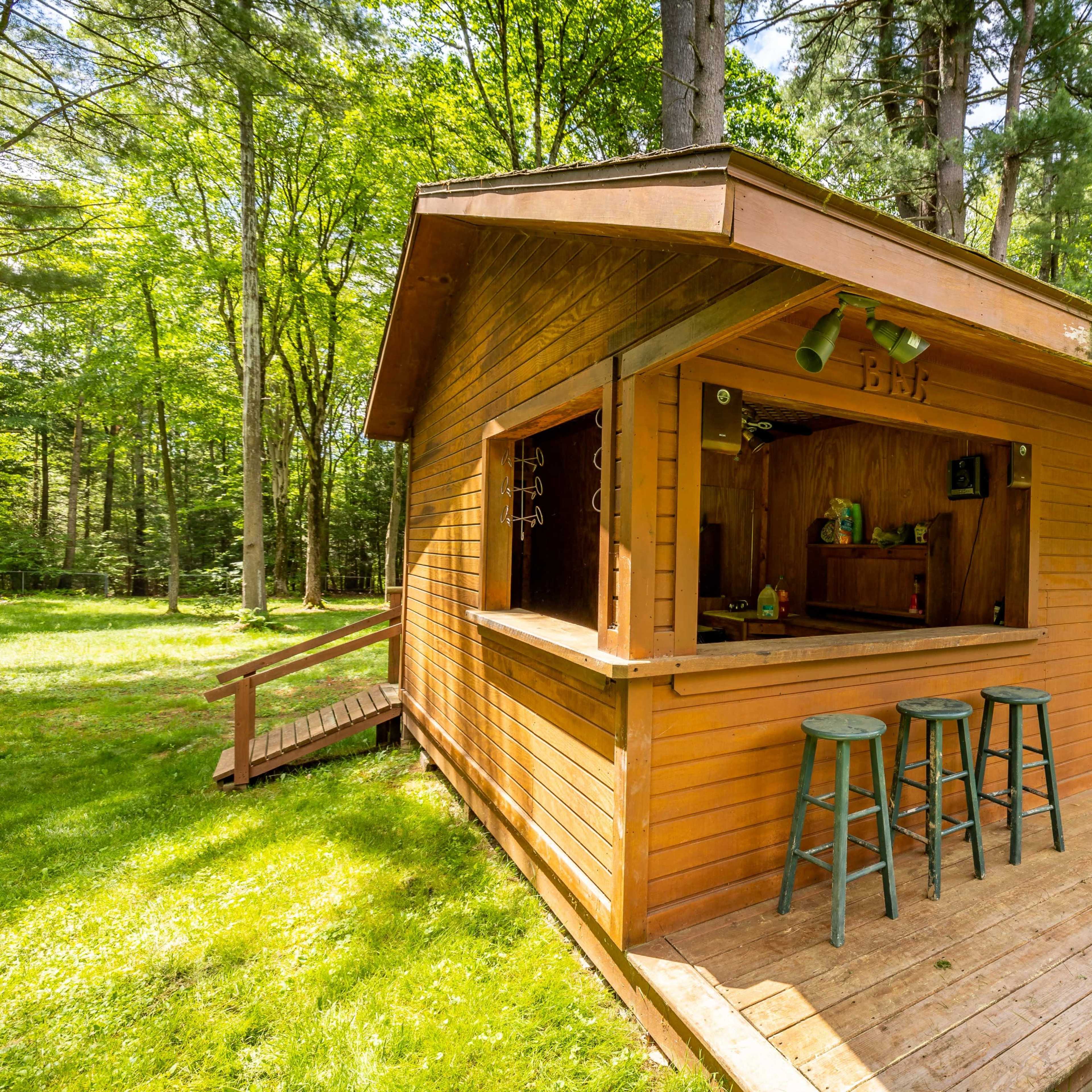 A wooden bar shed with three stools sits next to a swimming pool in a grassy area surrounded by trees.