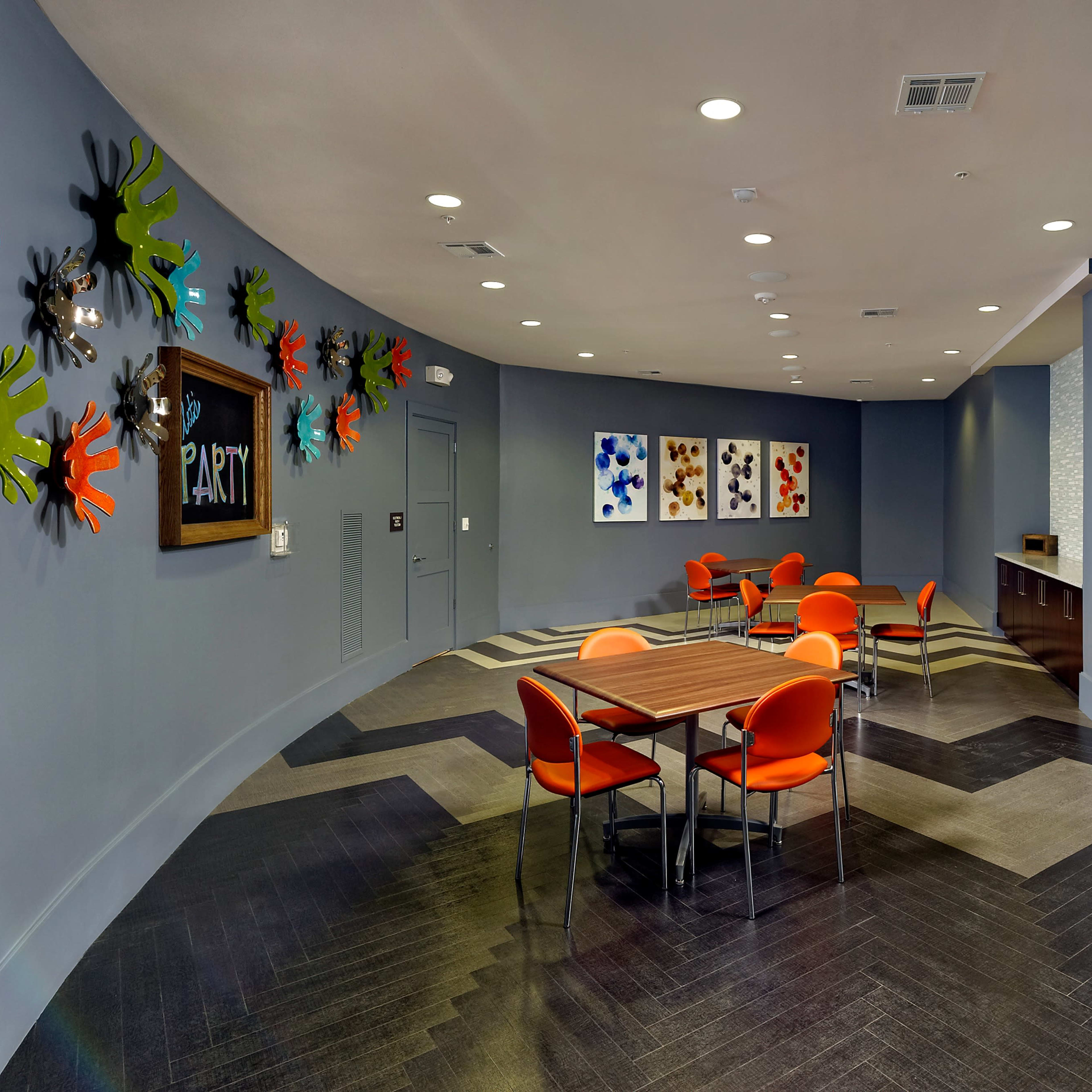 A brightly colored lounge area features orange chairs and a large table against a backdrop of playful wall art and a doorway to another room.