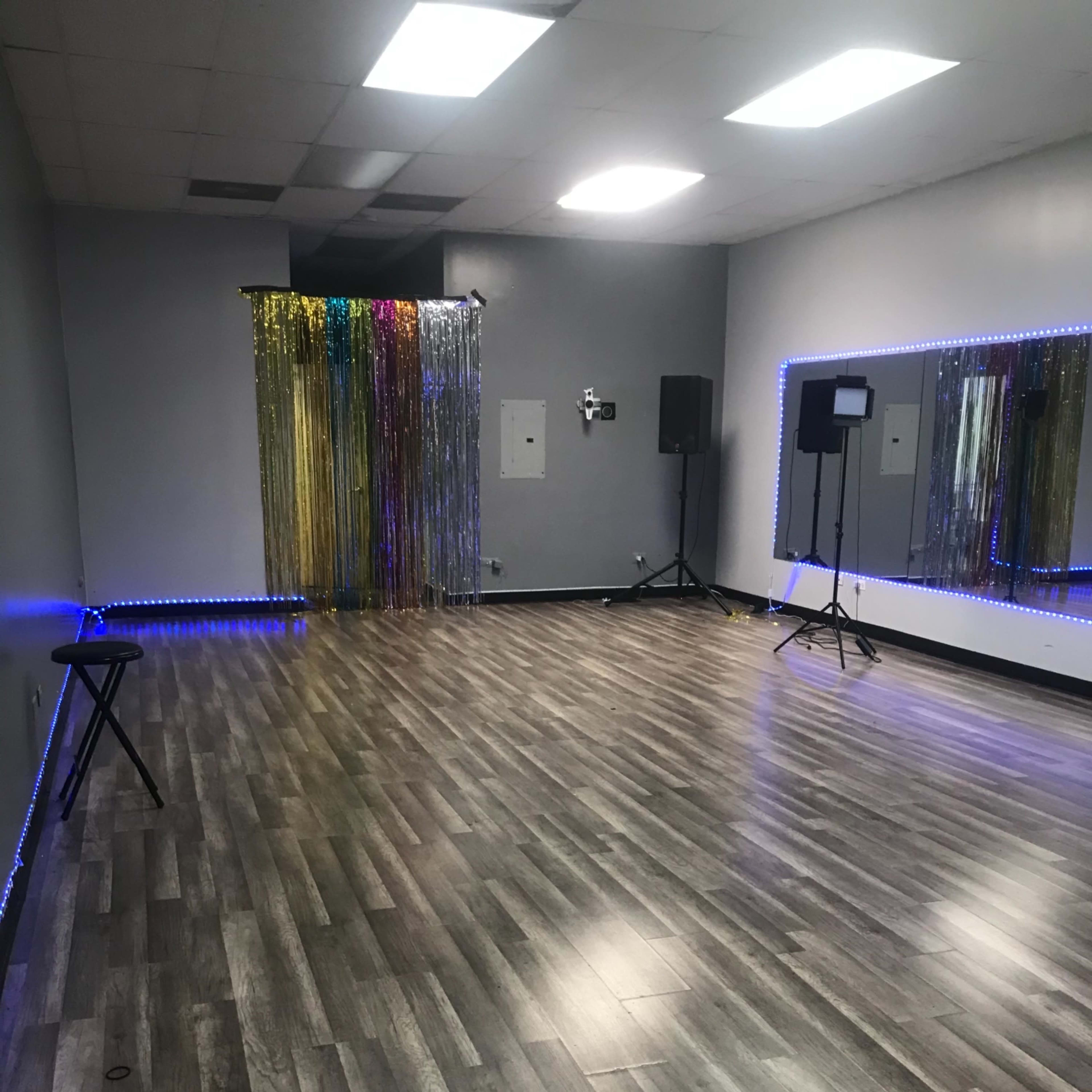 A spacious dance studio with wood-like flooring, a large mirror on one wall, and a backdrop of colorful streamers.