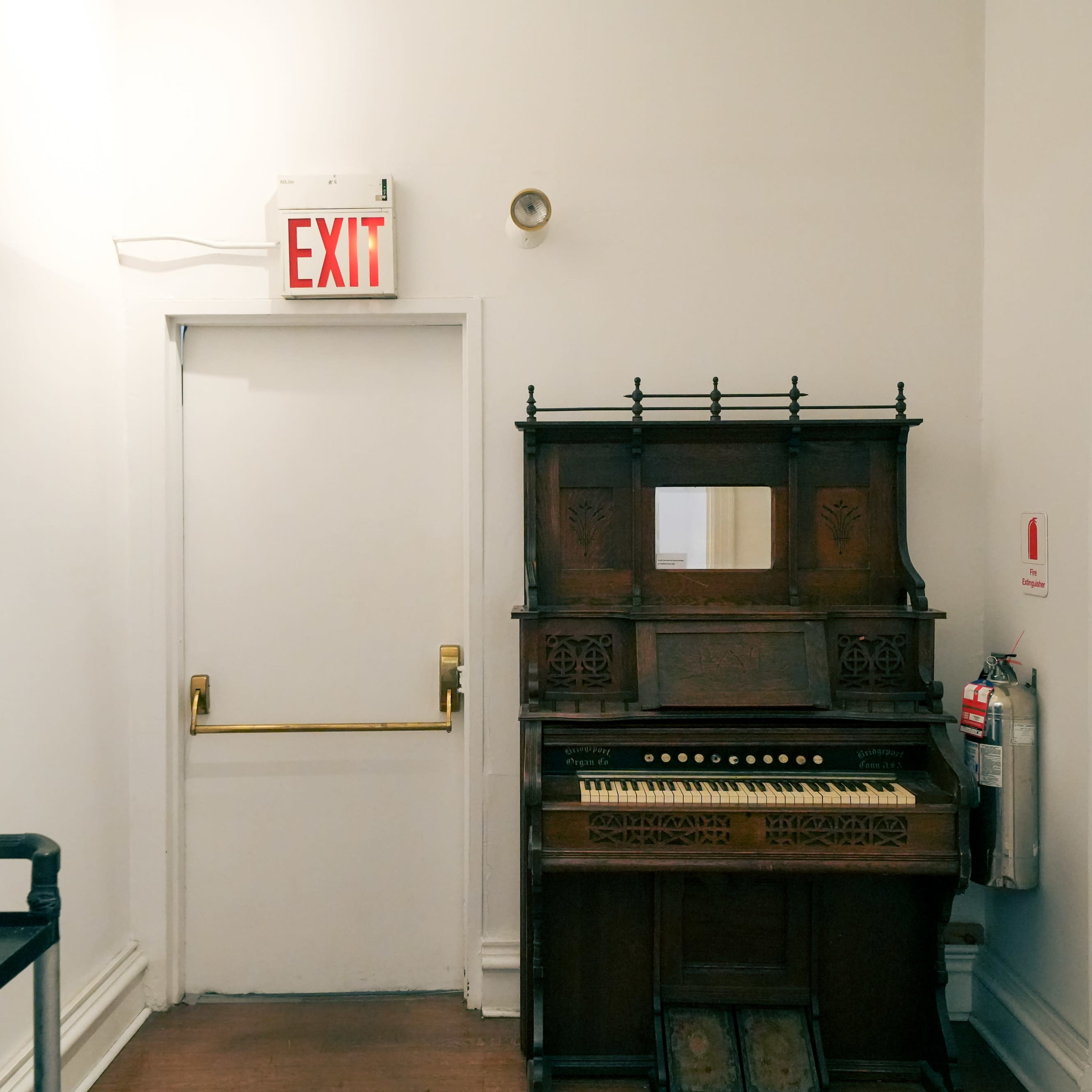 A vintage wooden piano positioned against a white wall near a door marked with an exit sign.