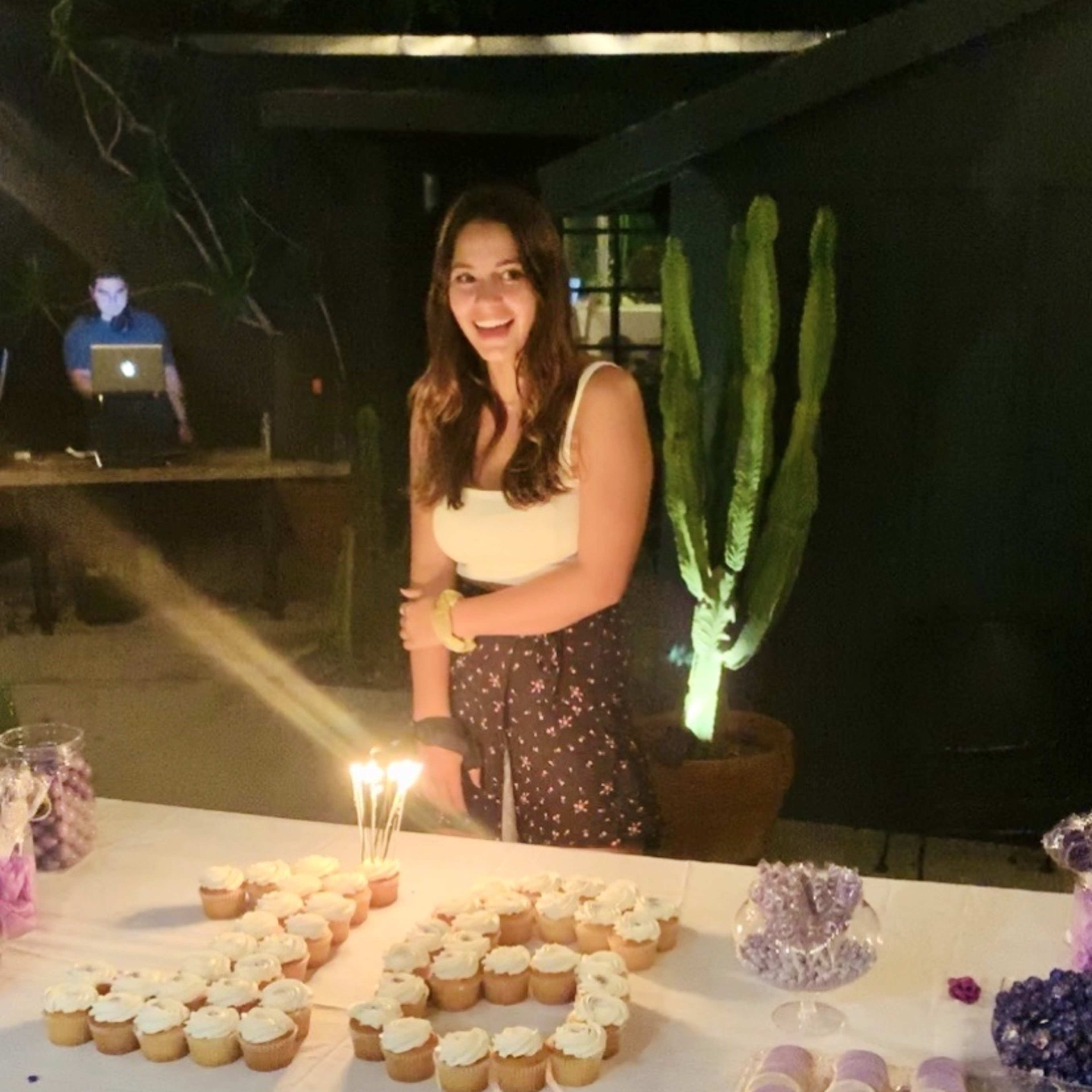 A woman stands beside a table decorated with cupcakes and a "30" made of cupcakes, with candles lit on top, in a dimly lit outdoor setting.