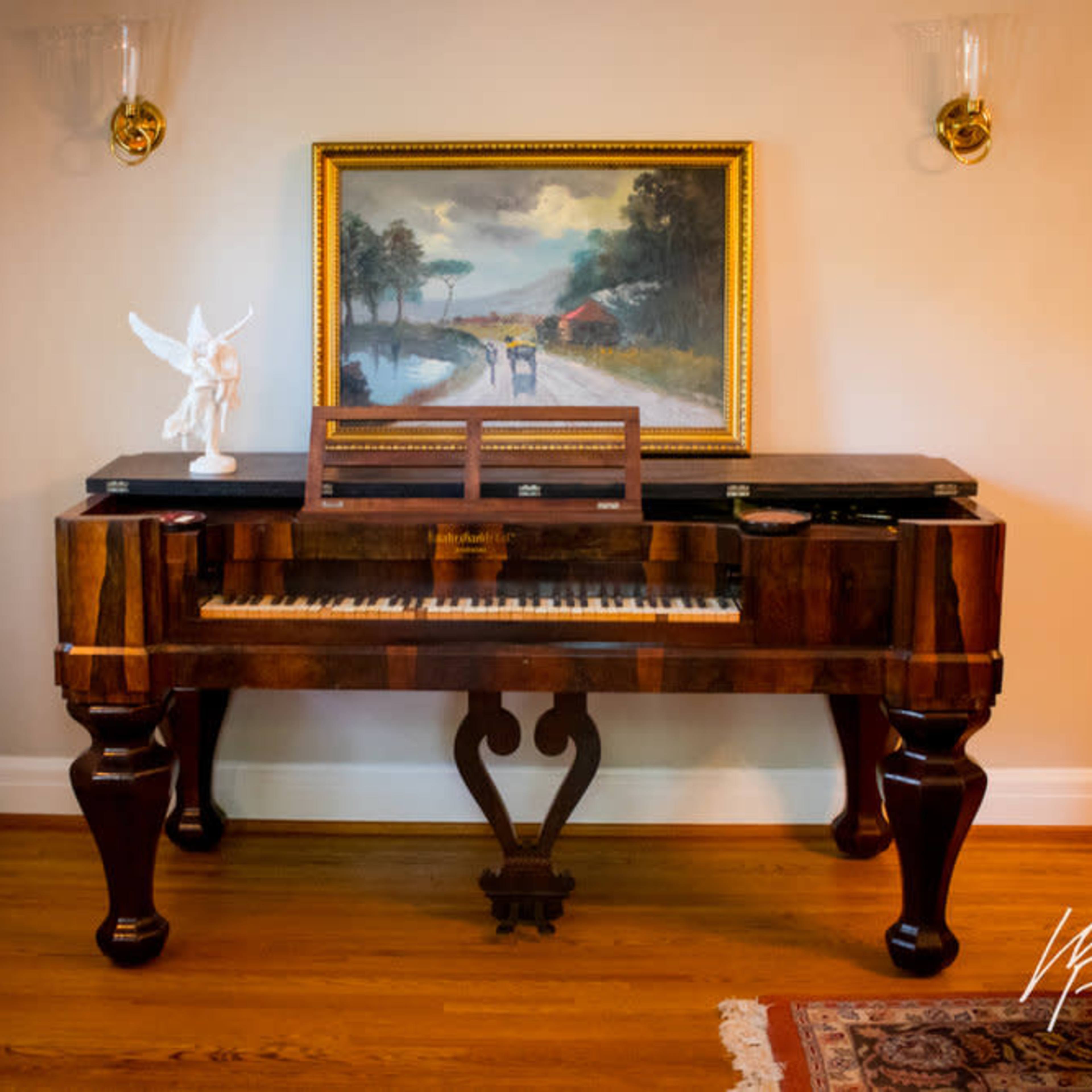 A wooden piano with decorative legs is positioned against a wall featuring a landscape painting and an angel figurine on top.
