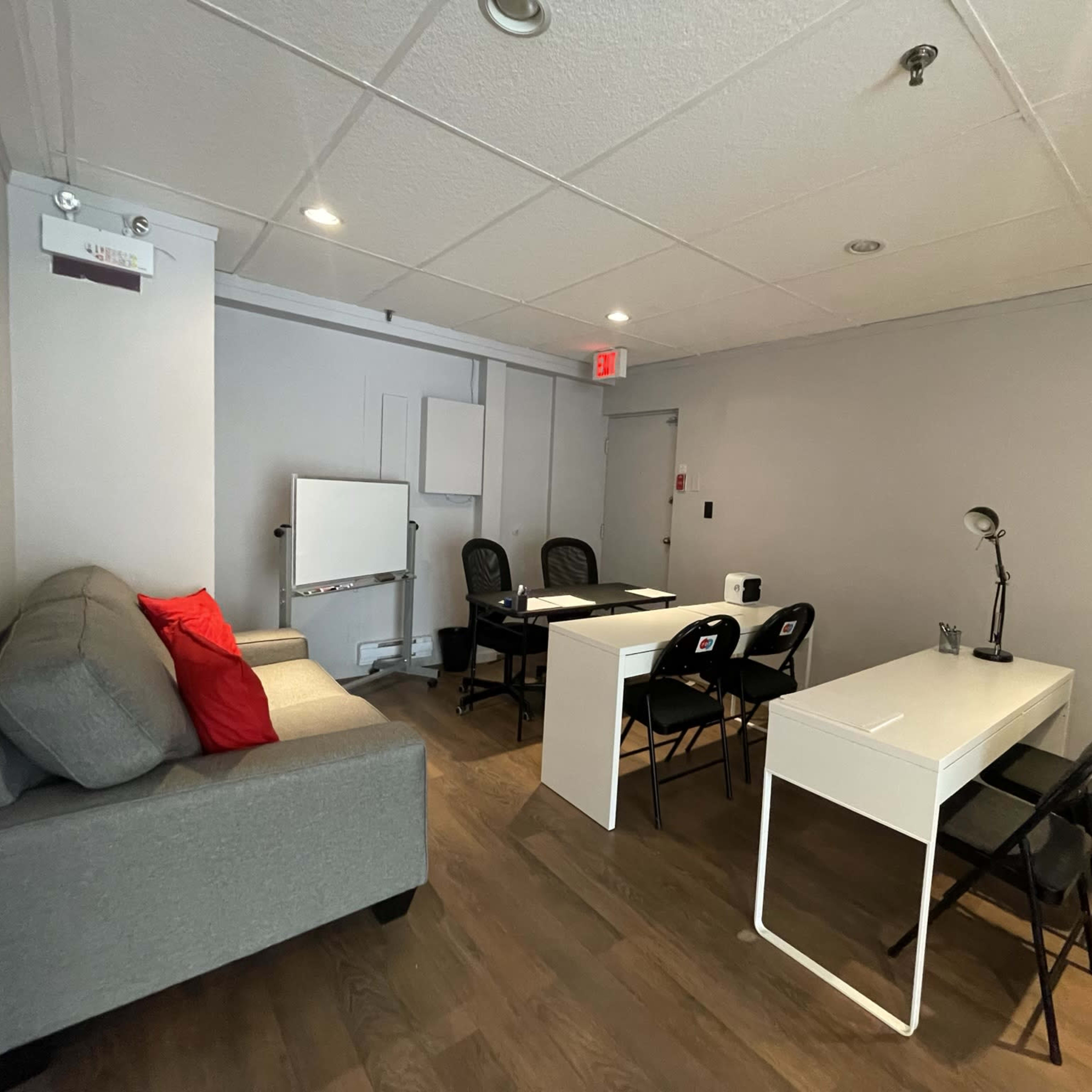 A modern office space features a gray sofa with a red cushion, two white desks with black chairs, and a whiteboard against the wall.
