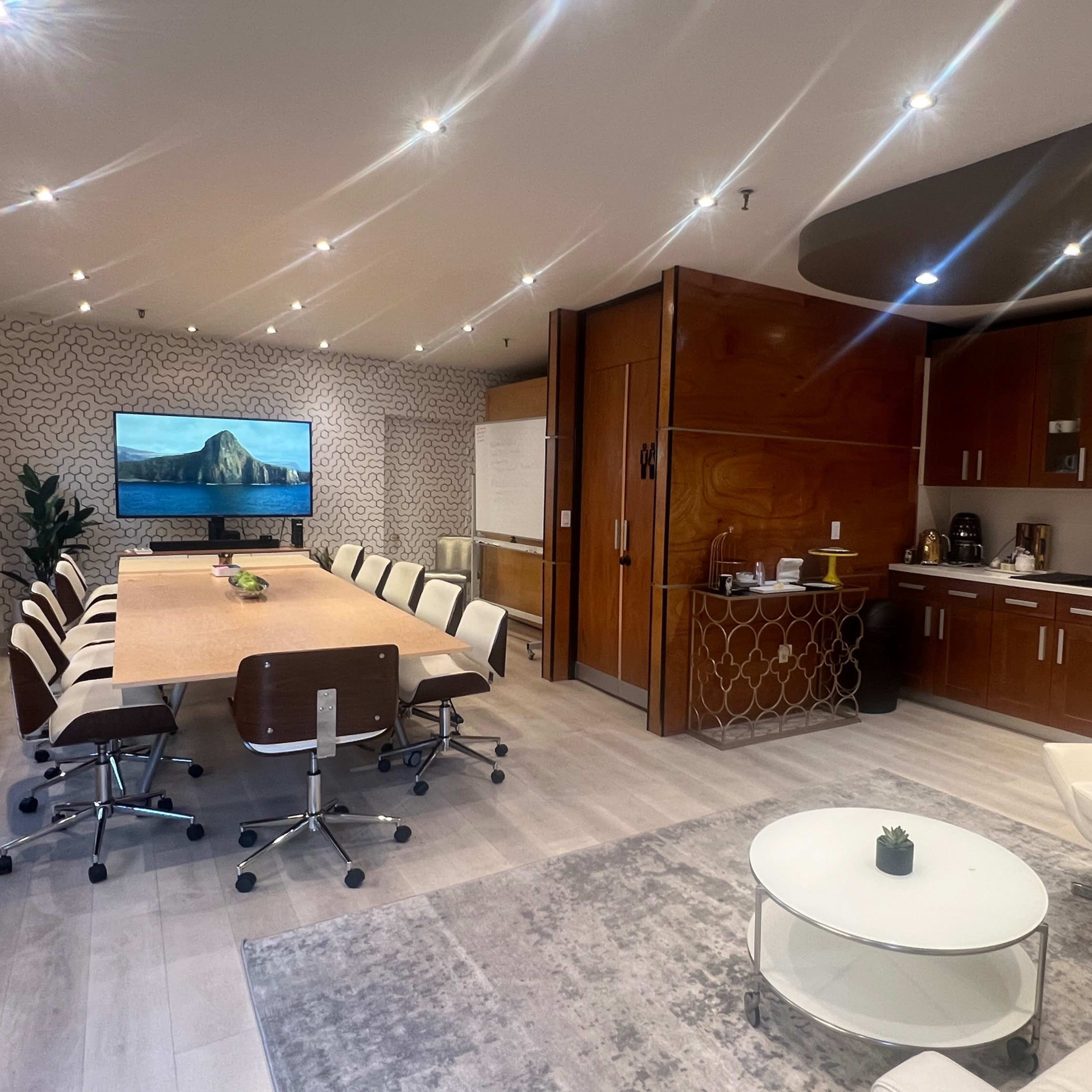 A modern conference room with a large table surrounded by chairs, a kitchenette, and a television displaying a scenic landscape.
