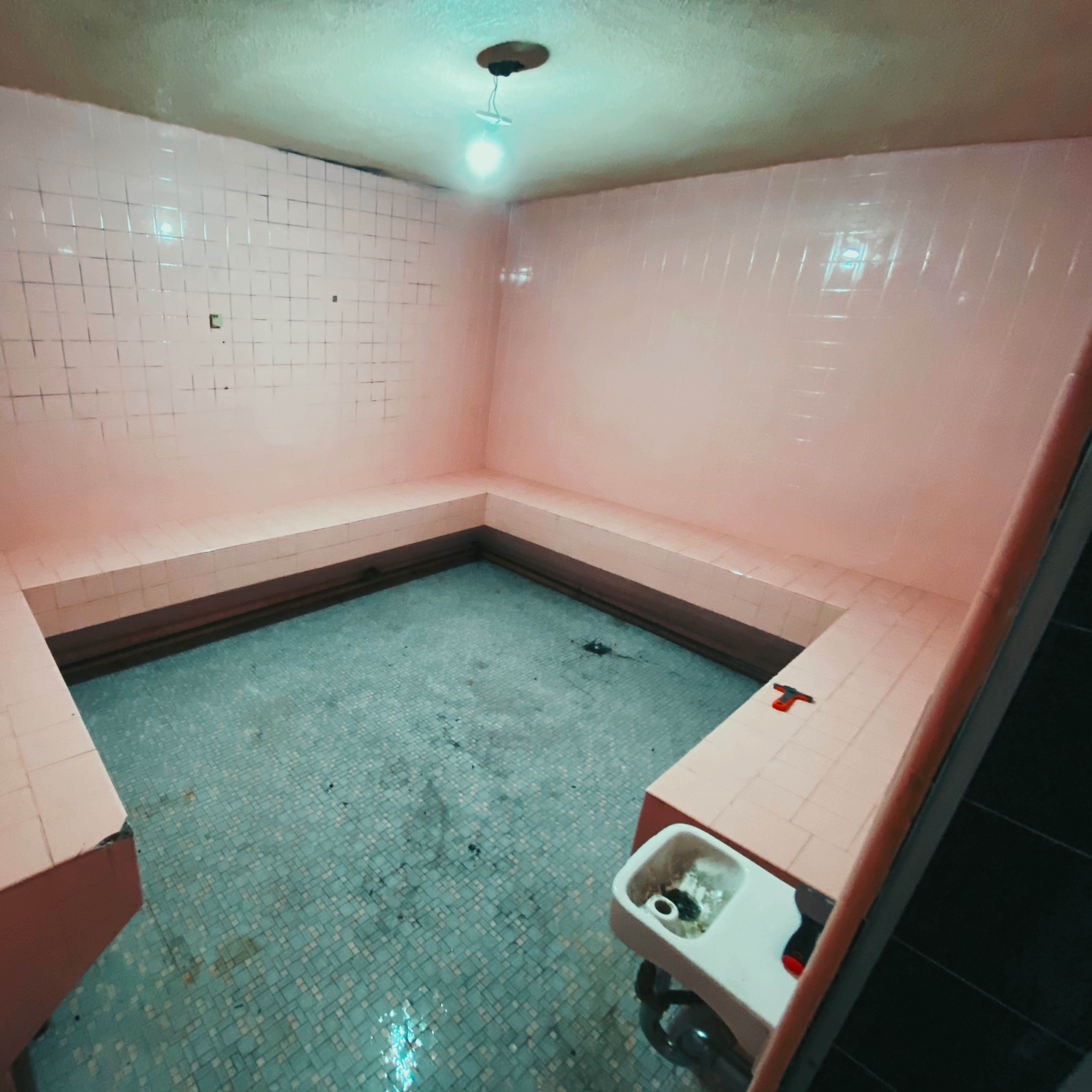 A small, empty room with pink tiled walls and a tiled floor featuring a drain and a sink in one corner.